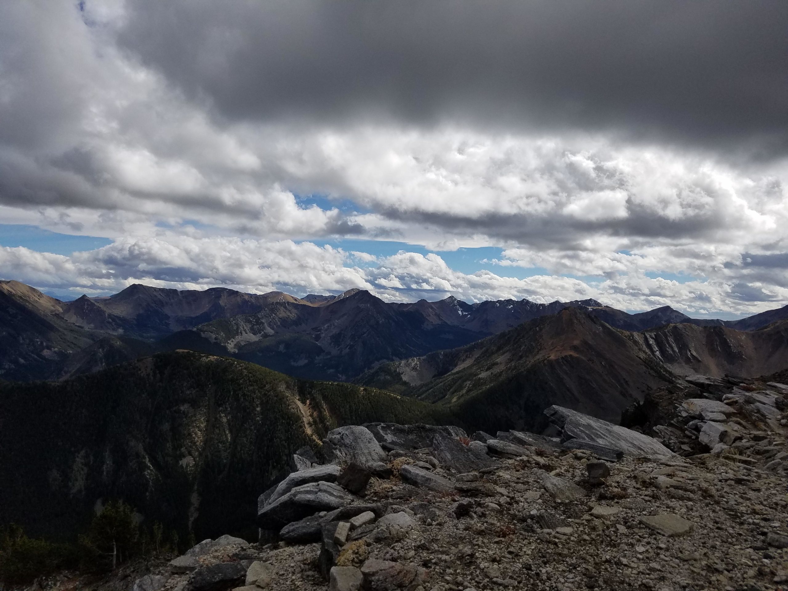 A panoramic view of rugged mountain peaks under a partly cloudy sky, showcasing a mix of green forests and rocky terrain. The foreground features stones and boulders, with distant mountains visible in varying shades of brown and gray. Curly Lake Highline mountain bike trail.