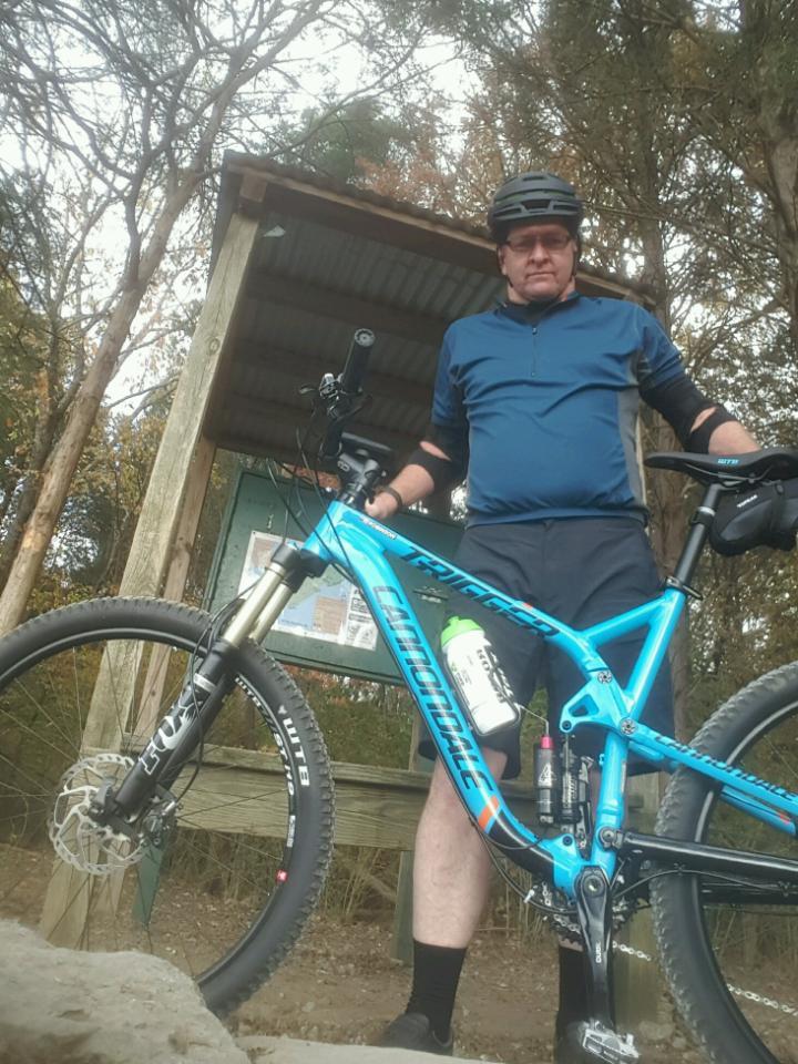Cannondale Trigger 27.5 3: A person in a blue cycling jersey and helmet stands next to a blue mountain bike at a trailhead, with wooden structures and trees in the background. The cyclist holds a water bottle in their bike frame and sports protective gear.