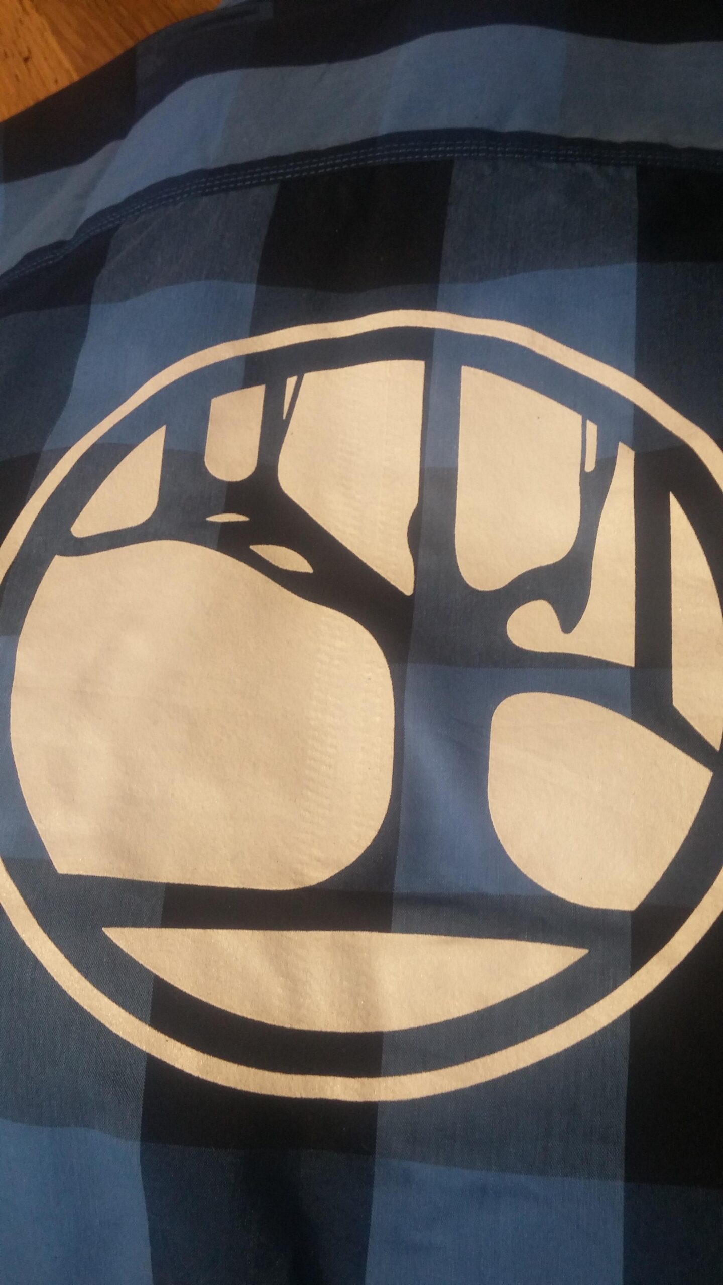 A close-up image of a blue plaid fabric featuring a large, circular logo in white. The logo includes a stylized tree design, with branches extending within the circle, contrasting against the blue checkered pattern of the fabric.