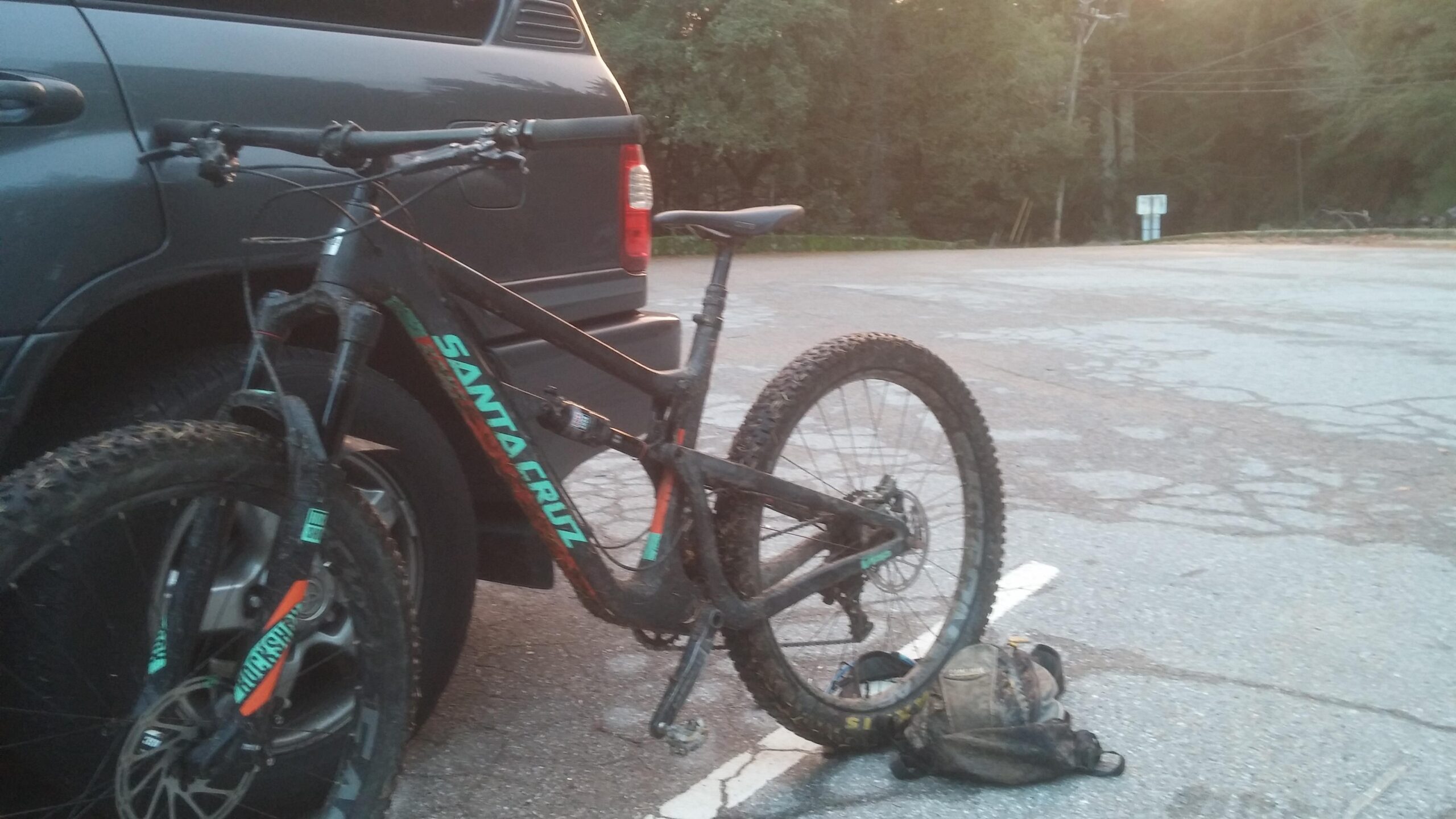 Santa Cruz Hightower 29er/27.5: A mountain bike leaning against a parked vehicle in a gravel parking lot. The bike has a black frame with colorful accents and muddy tires, indicating recent use. A small backpack is lying on the ground next to the bike, and the surrounding area is wooded with trees in the background.