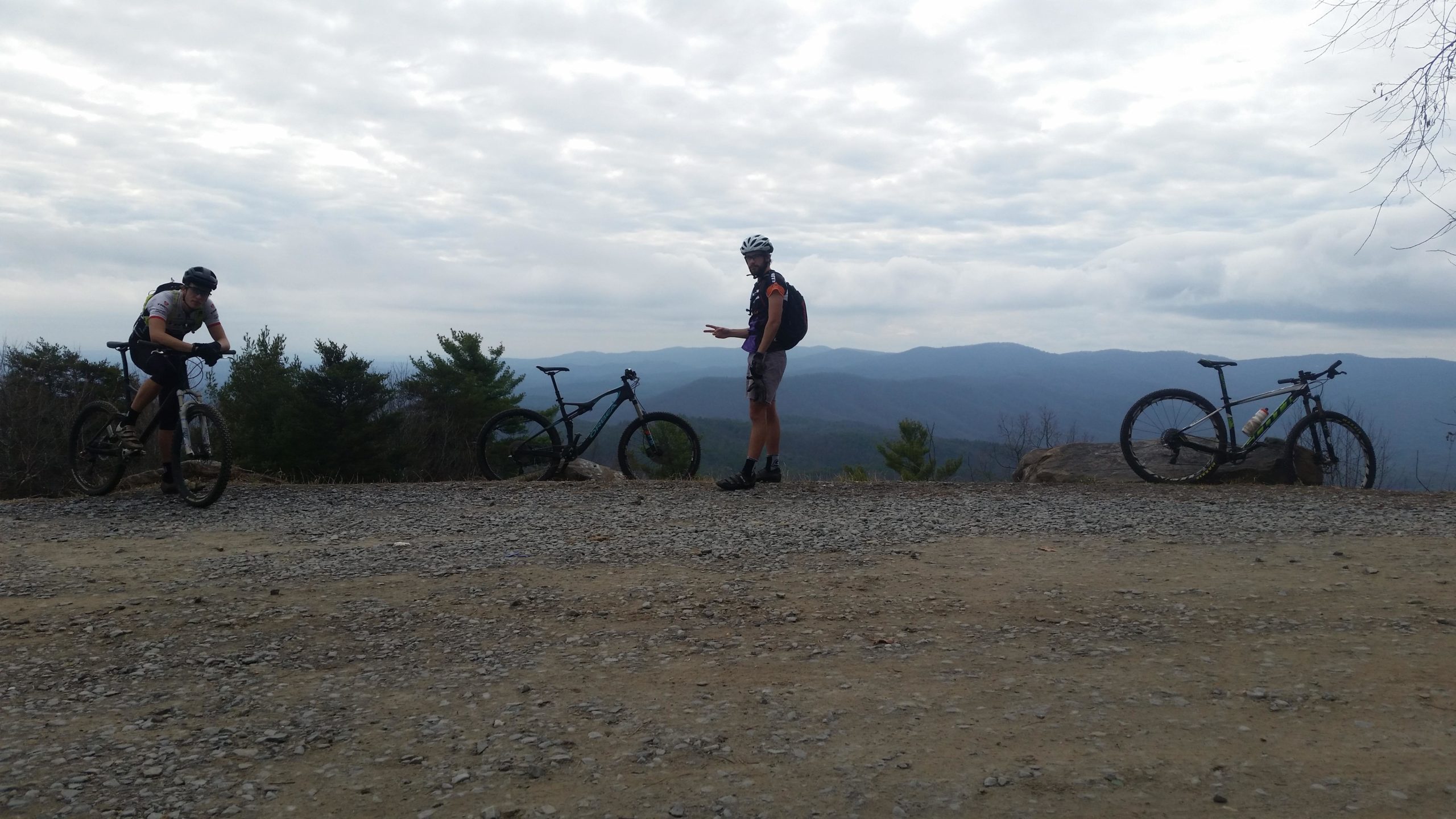 Two mountain bikers are seen on a gravel trail overlooking a scenic mountain view. One biker is seated on their bike, while the other stands nearby gesturing, both wearing helmets and athletic gear. In the background, a range of blue mountains is visible under a cloudy sky. Two bikes are parked beside them. Pinhoti Trail: Mountaintown Creek Segment mountain bike trail.