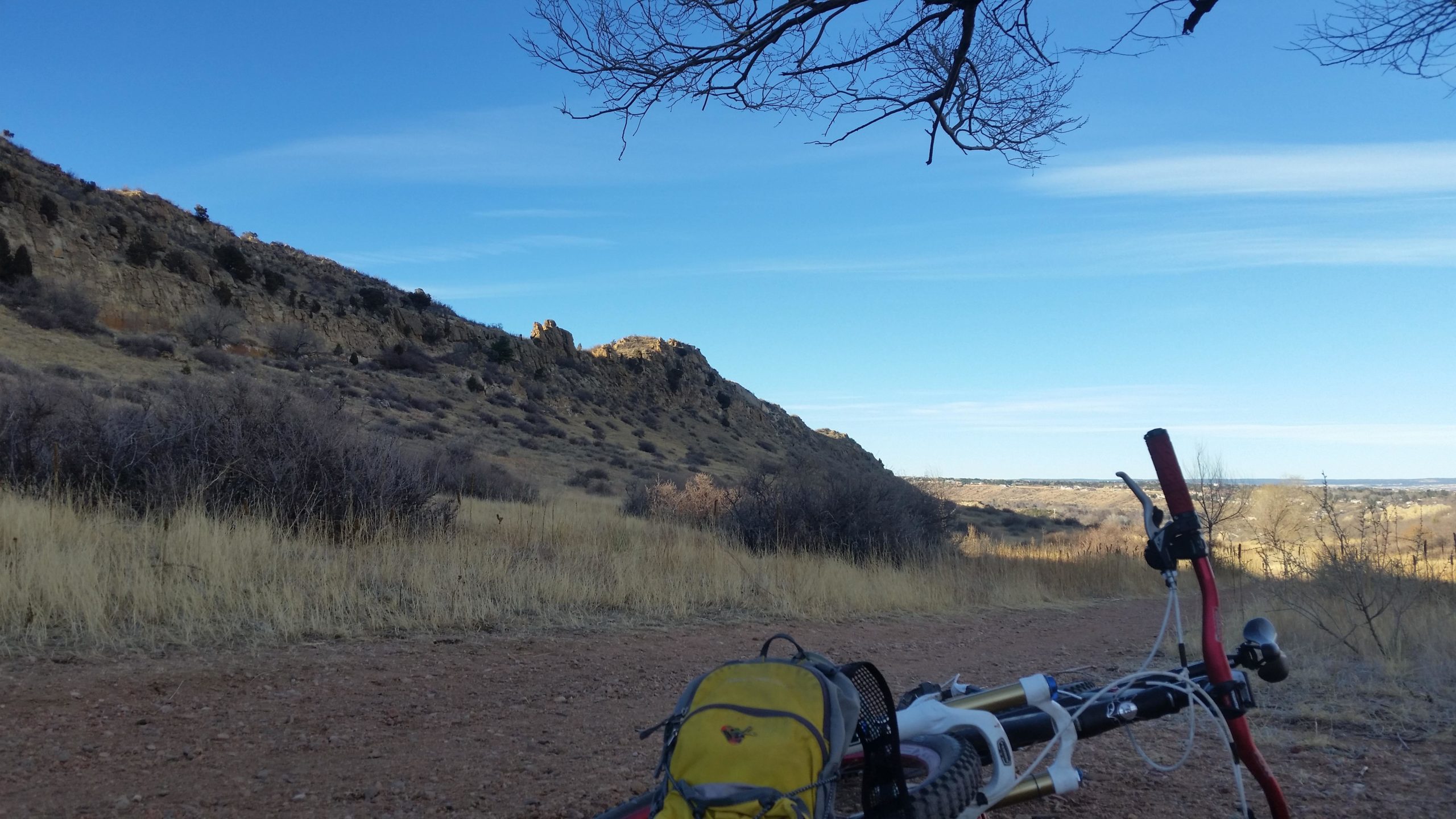 A scenic view of a hilly landscape with rugged terrain under a blue sky. In the foreground, a yellow backpack rests beside a mountain bike partially lying on a dirt path, with dry grass and shrubs surrounding the area. The hills in the background are dotted with trees and rocky outcrops, creating a serene outdoor atmosphere. Red Rock Canyon mountain bike trail.