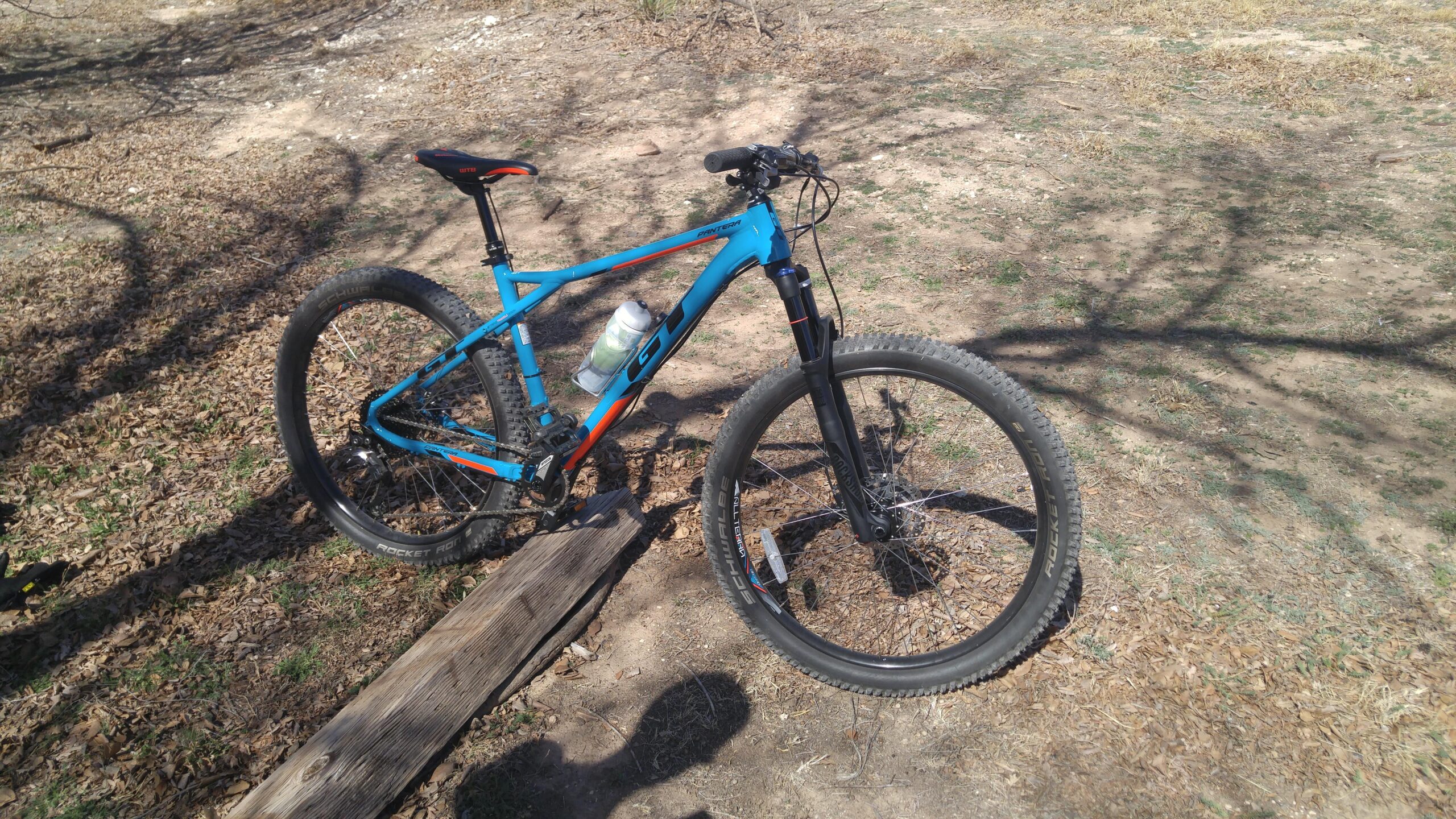 GT Pantera Expert: A bright blue mountain bike with orange accents is parked on a dirt trail surrounded by fallen leaves. The bike features a large front suspension fork and a water bottle attached to the frame. Shadows from nearby trees create patterns on the ground.