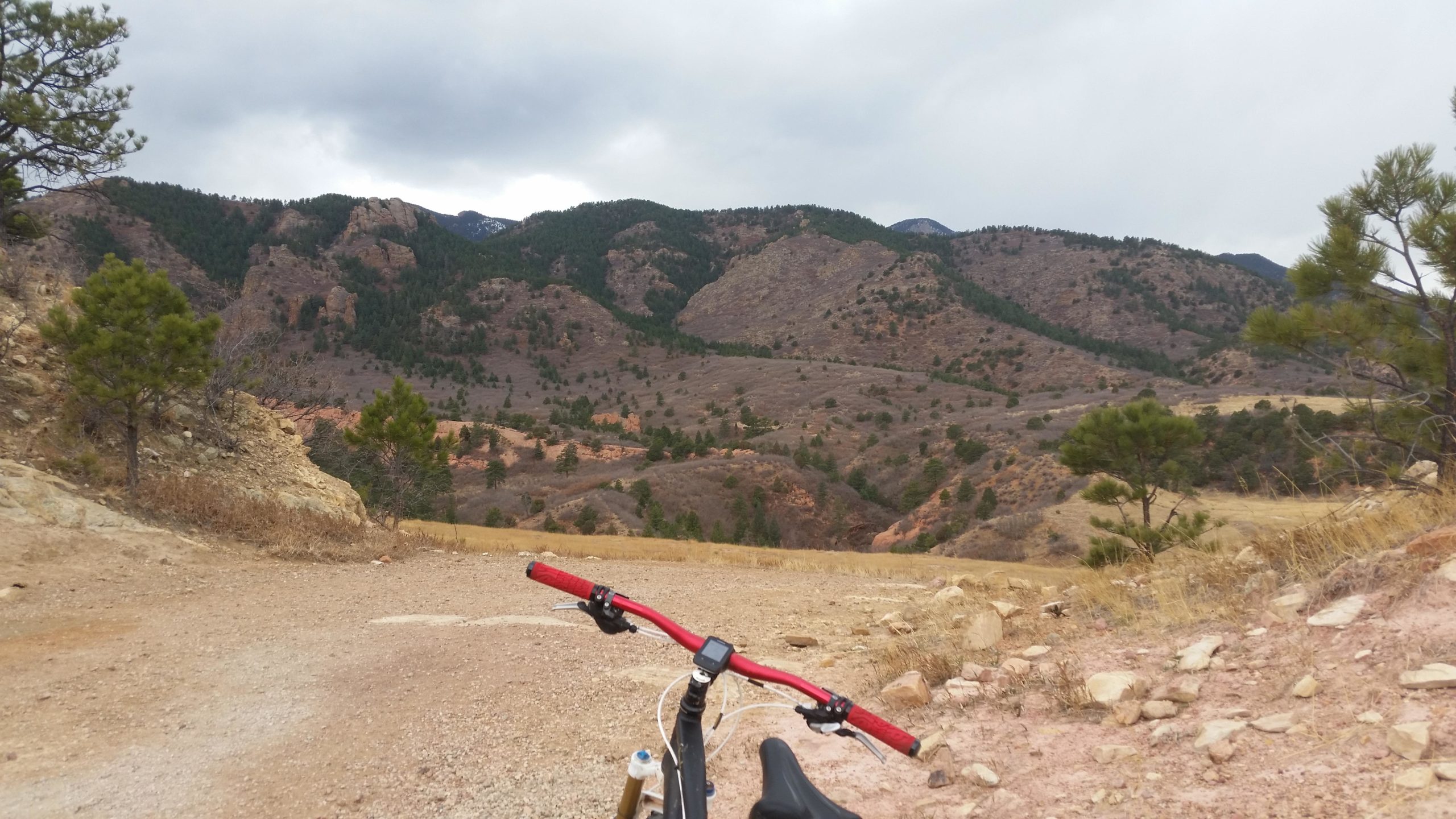 A mountain bike parked on a dirt trail, with a panoramic view of mountainous terrain and pine trees in the background under a cloudy sky. Red Rock Canyon mountain bike trail.