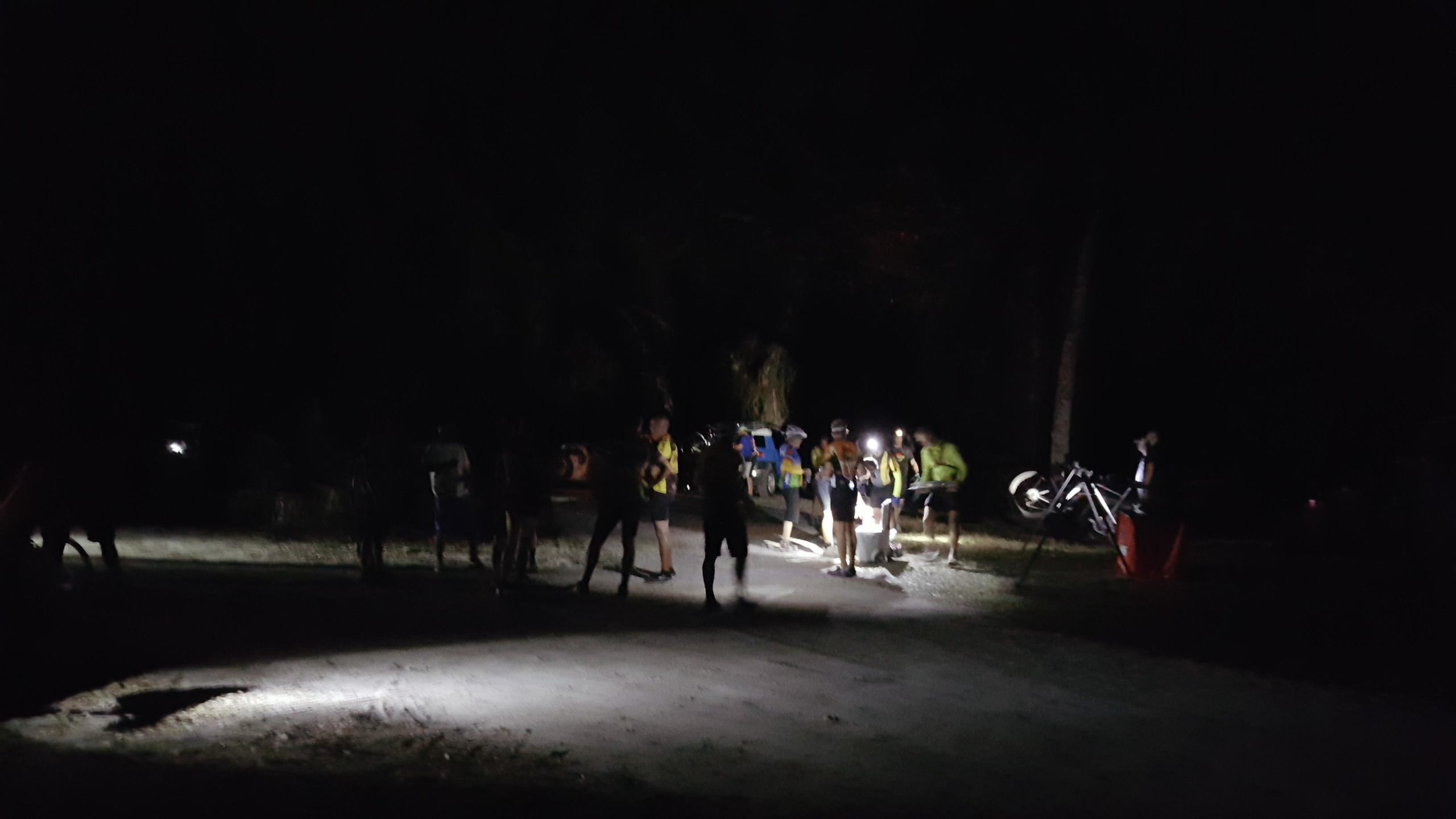 A group of people gathered at night on a gravel path, illuminated by headlamps and flashlights. Some individuals are standing together, while others appear to be checking equipment or discussing plans. A bicycle is leaning against a nearby post, adding to the outdoor activity scene. The background is mostly dark, with hints of vegetation and shadows surrounding the area. Carlton Preserve mountain bike trail.