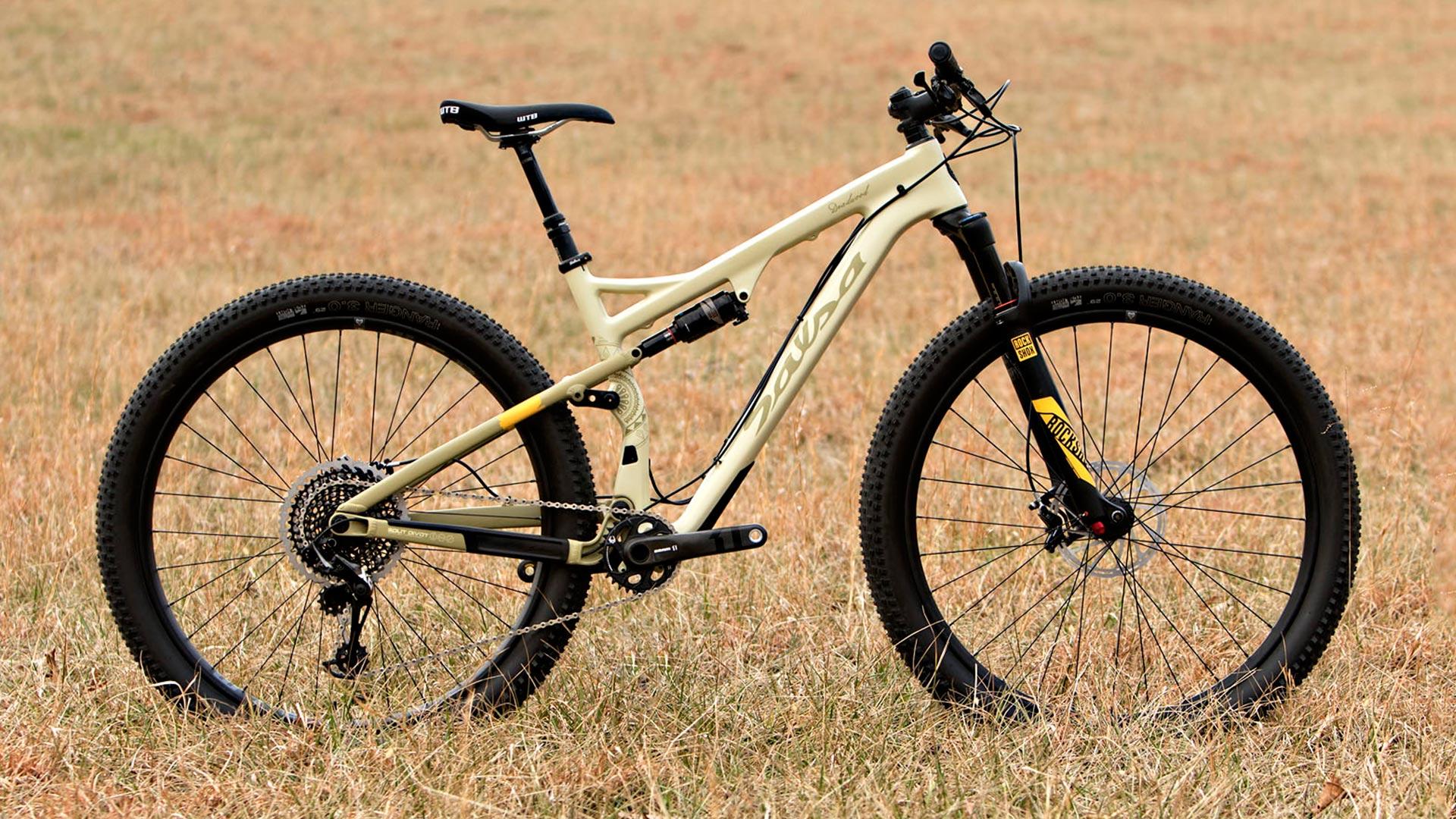 Salsa Deadwood Sus: A cream-colored mountain bike is positioned on dry, grassy terrain. The bike features thick black tires and a suspension fork, with prominent gears and a sleek frame design. The overall setup suggests it is built for off-road cycling.