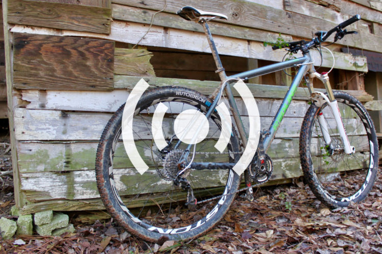A mountain bike with muddy tires is leaned against a weathered wooden wall, partially covered by peeling paint. The ground is littered with fallen leaves and small stones, creating a rustic outdoor setting.