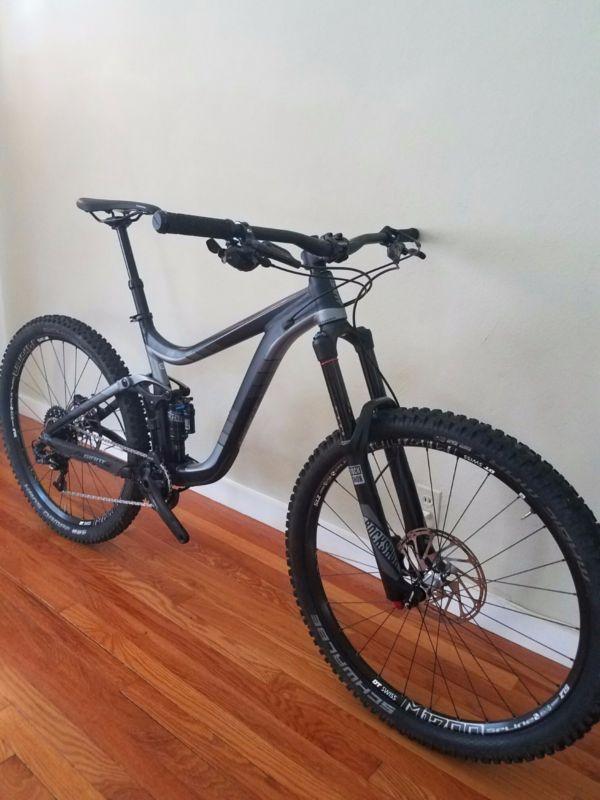 Giant Reign: A modern mountain bike with a sleek black frame, wide tires, and disc brakes, leaning against a light-colored wall on a wooden floor.