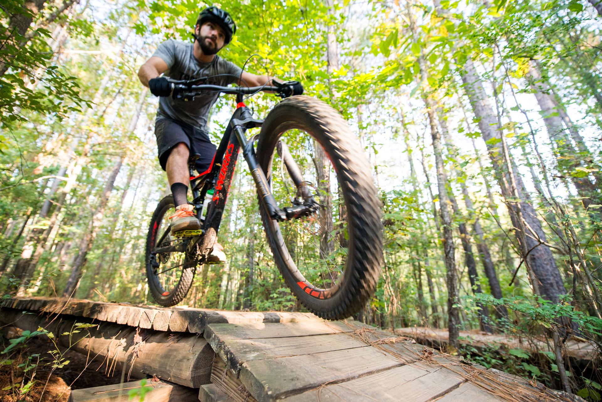 A mountain biker skillfully navigates a wooden bridge along a forest trail, surrounded by lush greenery and tall trees. The rider is in mid-motion, showcasing the bike's large tires as they soar over the bridge. USNWC mountain bike trail.