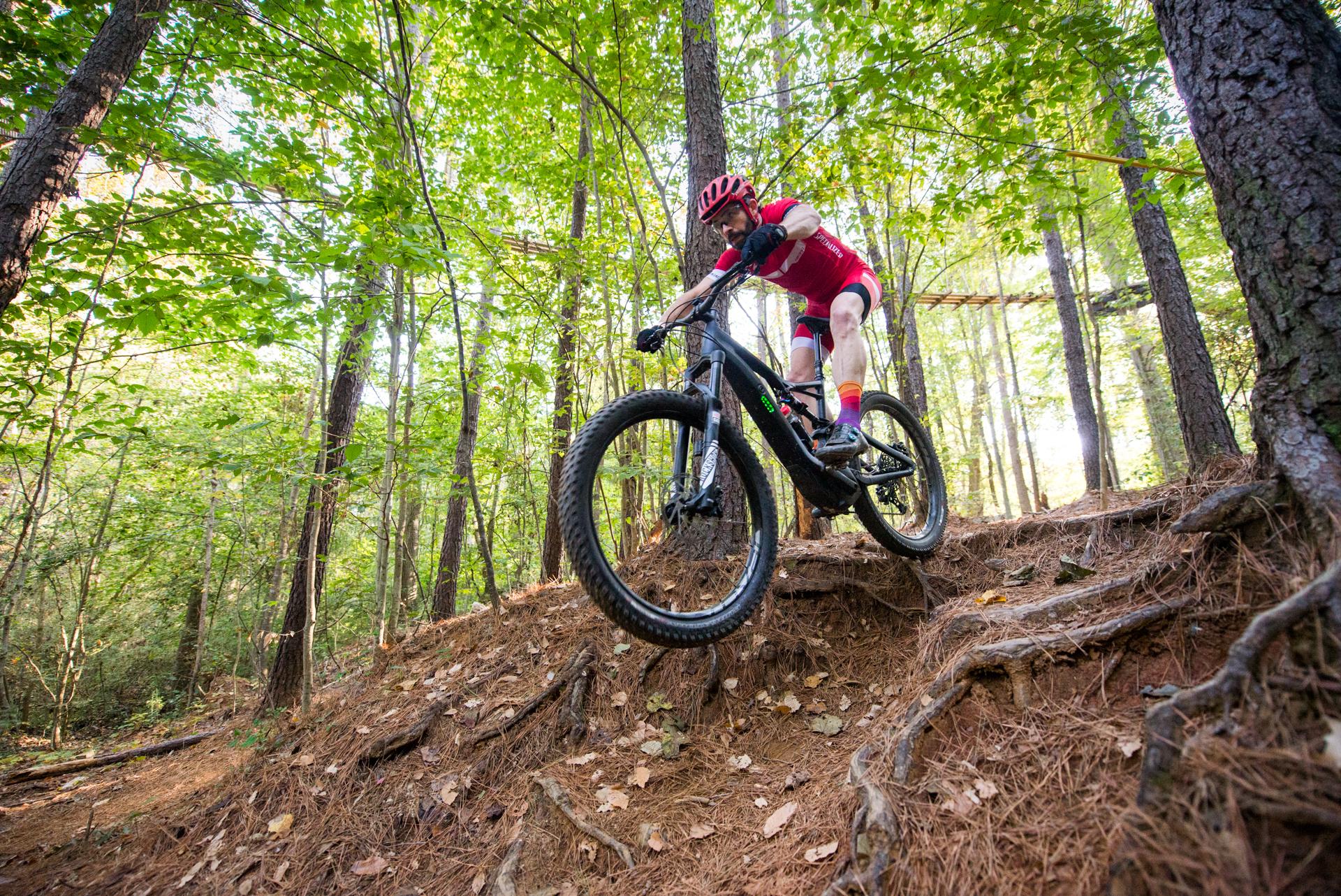 A mountain biker in a red outfit leaps over a root-covered incline on a trail surrounded by lush green trees. The sunlight filters through the leaves, creating a dynamic and energetic atmosphere. USNWC mountain bike trail.