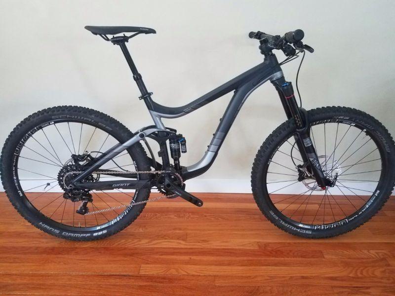 Giant Reign: A modern mountain bike with a sleek black and gray frame, featuring thick tires designed for off-road terrain. The bike has a full suspension system, visible rear shock, and a wide handlebar. It is positioned upright against a light-colored wall, with wooden flooring in the background.