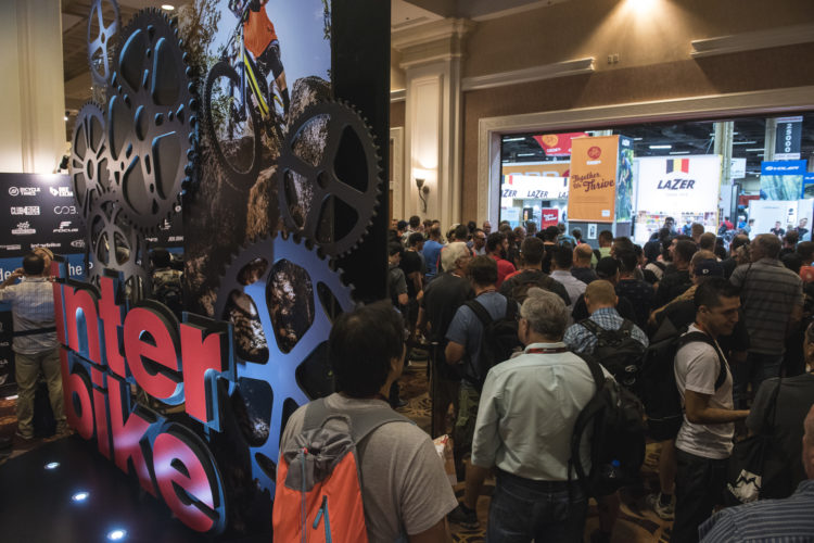 A crowded exhibition hall at the Interbike event, featuring a prominent display with large gear motifs and a mountain biker image. Attendees are gathered near the entrance, showcasing the energy and excitement of the cycling industry gathering.