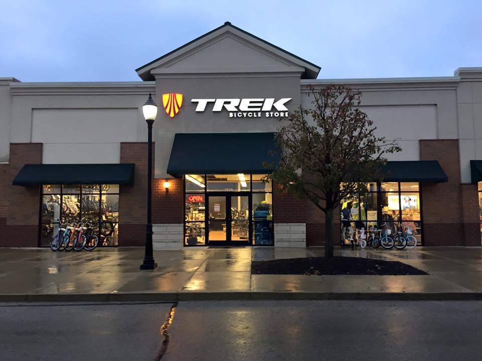 Exterior view of a Trek Bicycle Store, featuring large windows displaying various bicycles. The store's name and logo are prominently displayed above the entrance, and a lamppost stands nearby. The setting appears to be in a shopping plaza during twilight.