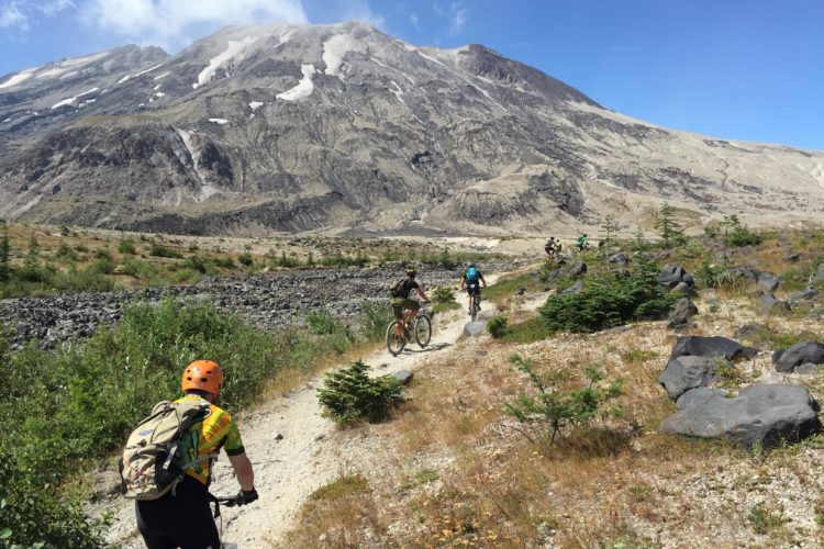 Mountain bikers riding on a dirt trail surrounded by rocky terrain and sparse vegetation, with a towering mountain in the background under a clear blue sky.