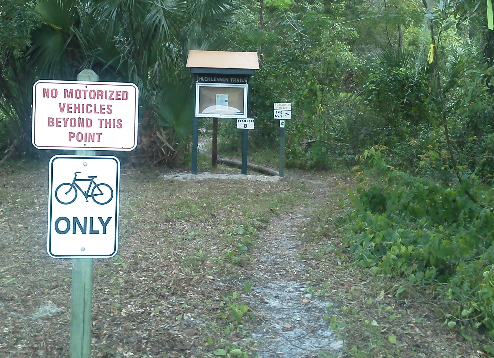 Two signs are visible at the entrance of a trail. The first sign, which is larger and prominently displayed, reads "No Motorized Vehicles Beyond This Point." Below it, a smaller sign indicates "Bicycles Only." In the background, there is a wooden information board labeled "Chuck Lennon Trails," with various trail markers nearby. The area is surrounded by lush greenery and a dirt path leads further into the trail. Chuck Lennon Park mountain bike trail.