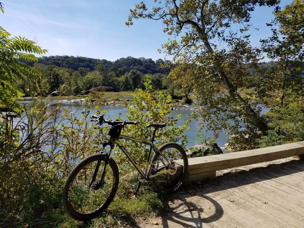 Giant Talon 27.5 4: A mountain bike resting near a riverbank, surrounded by greenery and trees, with sunlit hills in the background under a clear blue sky.