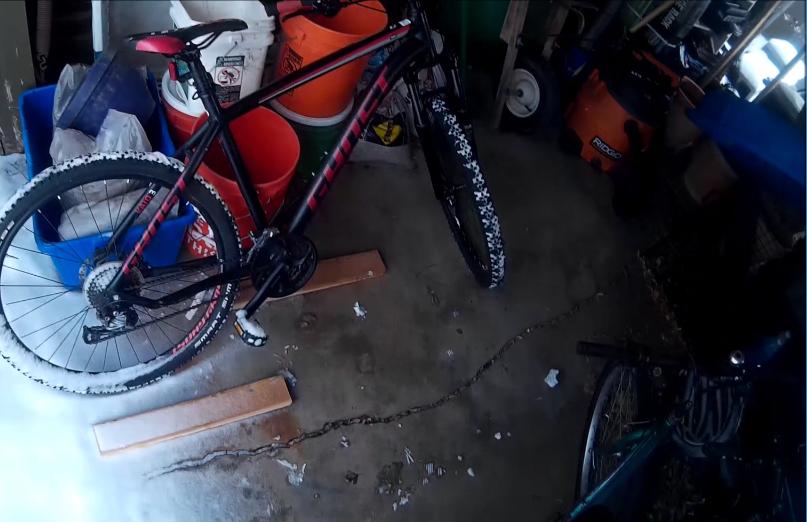 Ghost Kato 3: A black mountain bike with red accents is leaning against wooden planks in a cluttered garage. In the background, there are buckets, a vacuum, and various items stacked in disarray. The floor is concrete with some scattered debris.