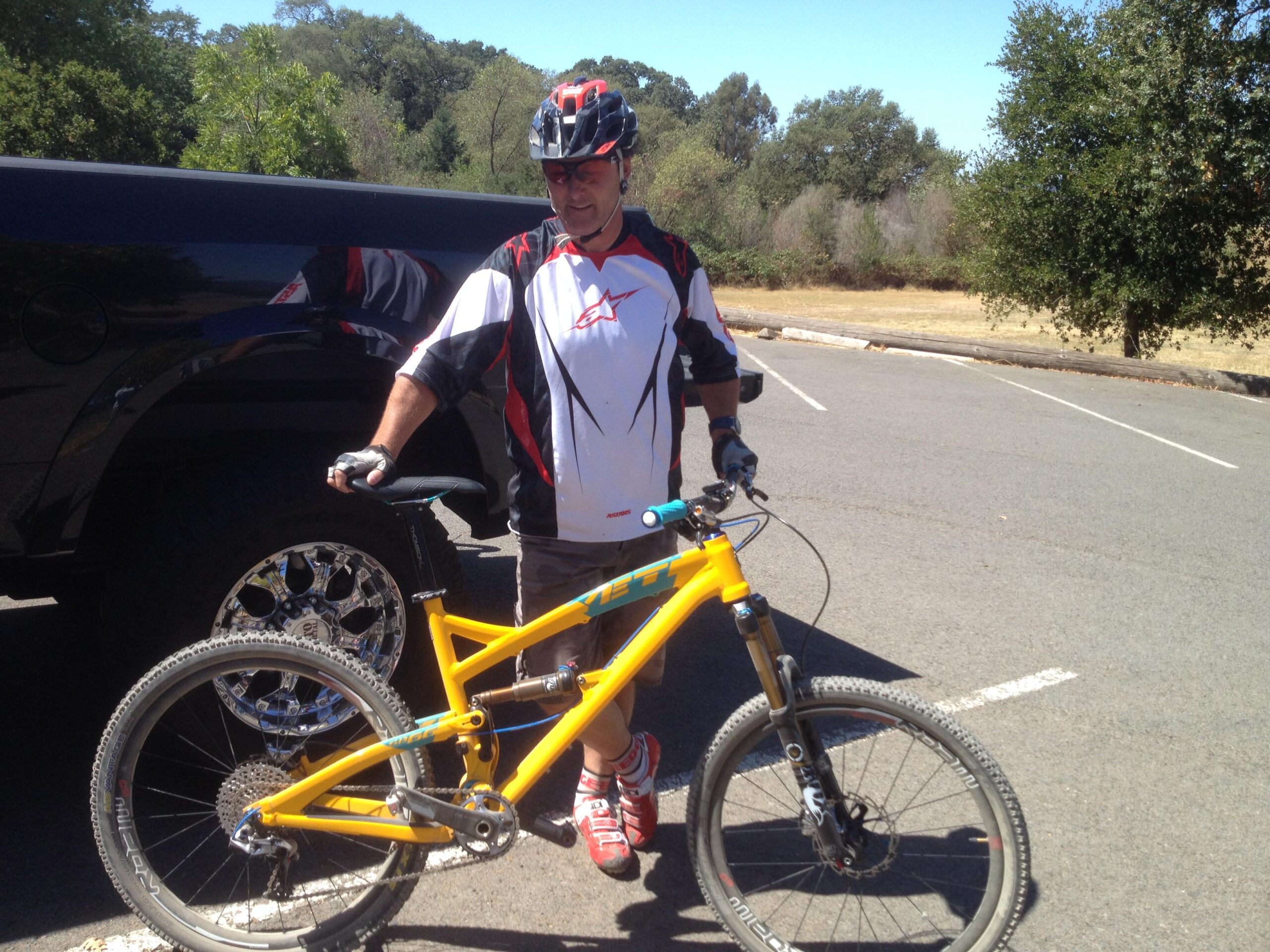 Yeti SB66: A cyclist in a helmet and sportswear stands next to a bright yellow mountain bike parked beside a black vehicle. The setting appears to be an outdoor area with trees in the background and a paved lot.
