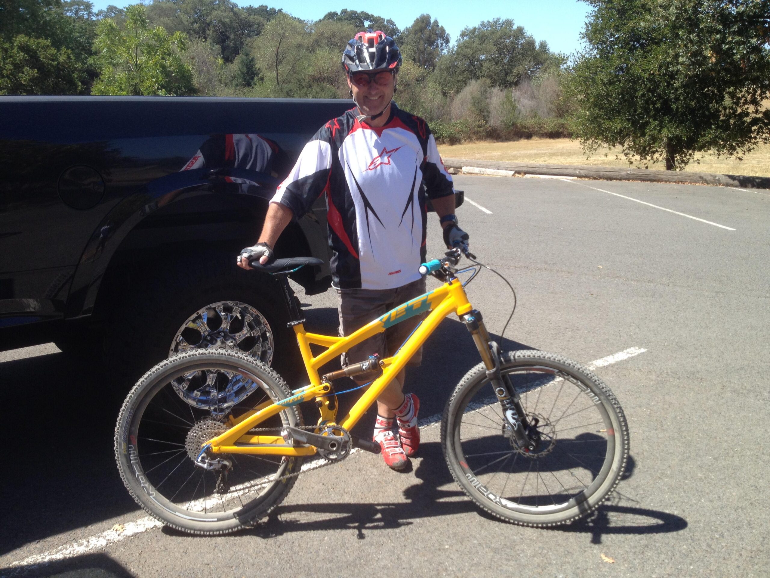 Yeti SB66: A person in a cycling outfit stands next to a bright yellow mountain bike, smiling. They are wearing a helmet and gloves, and are positioned in a parking area with a black vehicle behind them. The background features trees and open spaces, indicating an outdoor location.