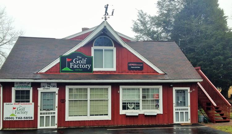 A red wooden building with a peaked roof, featuring a sign that reads "The Golf Factory." The façade has multiple windows and a door, along with a small staircase leading to a second level. A weather vane is visible at the top of the roof. Trees are in the background, and the scene appears overcast and rainy.