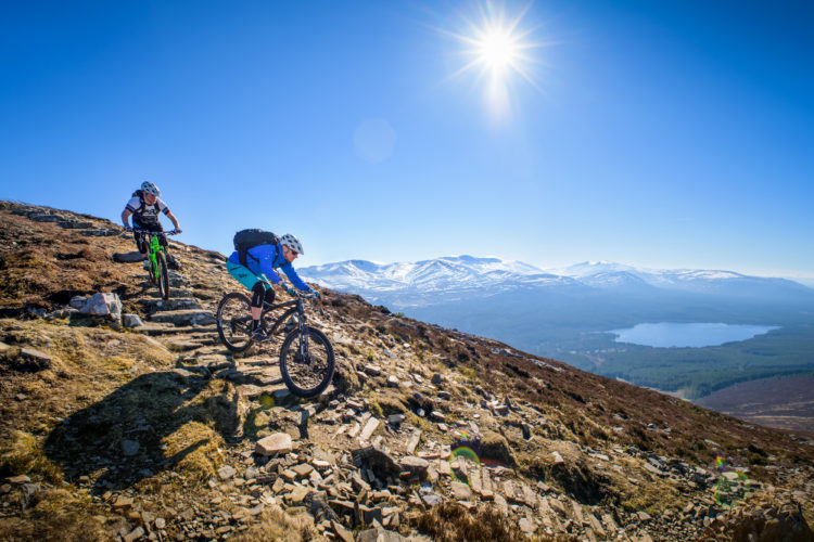Two mountain bikers navigate a rocky trail on a sunny day, surrounded by a stunning landscape of mountains and a lake in the distance. The sky is clear blue, and snow-capped peaks are visible in the background. One rider is in the foreground, focused on maneuvering their bike, while the other follows closely behind.