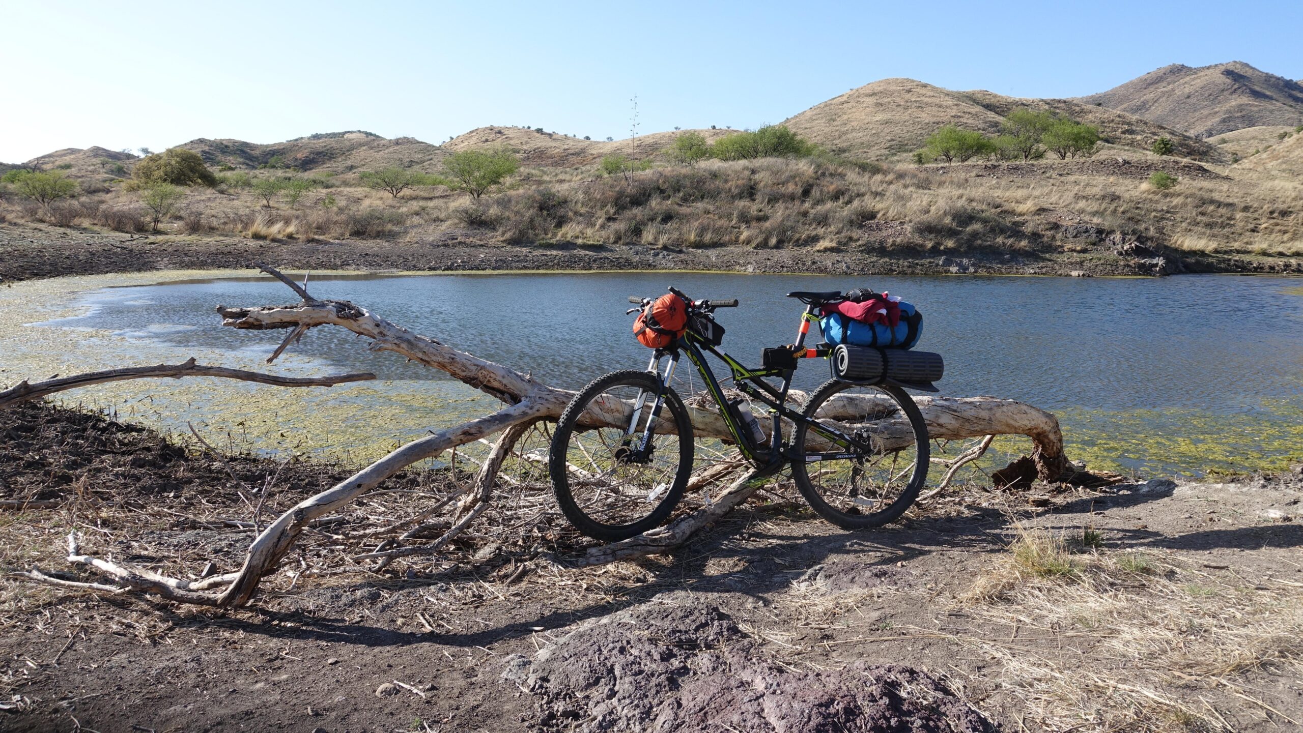 Specialized Camber: A mountain bike equipped with colorful bags is parked beside a small lake, surrounded by dry hills and vegetation. A fallen log stretches across the foreground, and the water reflects the clear blue sky.