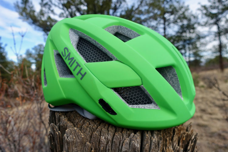 A vibrant green bicycle helmet resting on a wooden stump, with a blurred natural background of trees and shrubs. The helmet features ventilation openings and branding on the side.