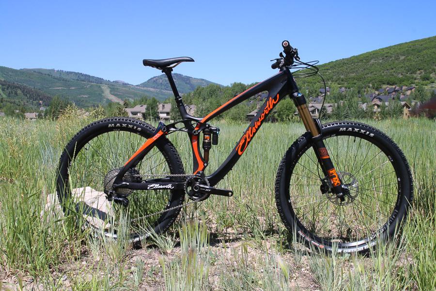 Ellsworth Rogue Forty: A mountain bike with an orange and black design stands on rocky ground amidst tall grass, surrounded by green hills and a clear blue sky.