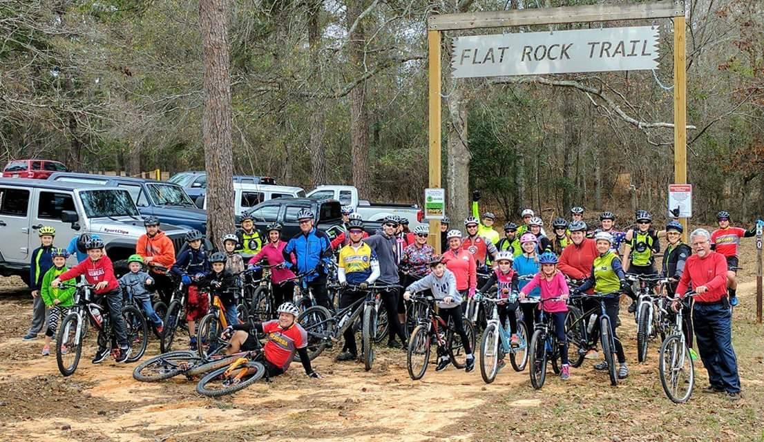 A large group of cyclists, including children and adults, gather at the entrance of the Flat Rock Trail. They are posing with their bikes in an outdoor setting surrounded by trees and parked vehicles in the background. The trail sign is prominently displayed overhead. Flat Rock Park mountain bike trail.