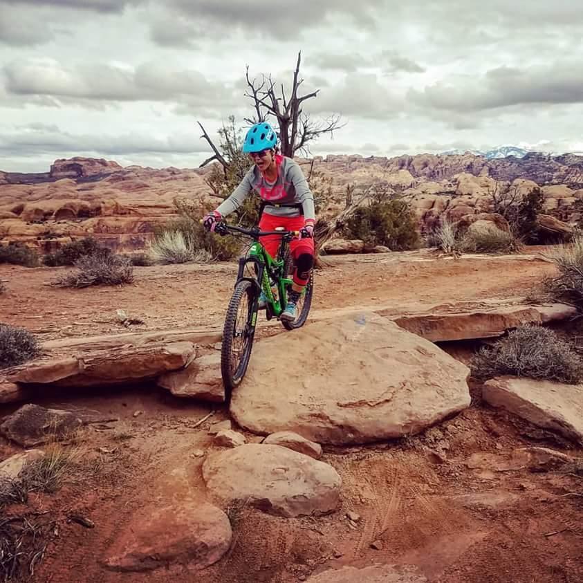 A mountain biker in vibrant gear navigates over a rocky terrain, riding a green bike. The landscape features rugged stone formations and sparse vegetation under a cloudy sky. Hymasa mountain bike trail.
