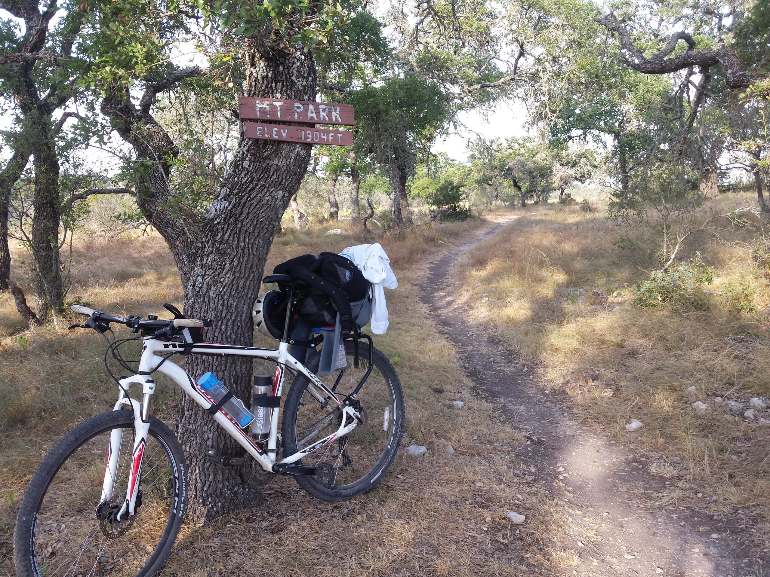 A mountain bike resting against a tree with a sign that reads "Mt. Park Elev. 1904 ft." The background features a dirt path winding through a grassy area with scattered trees. The bike has a water bottle and a helmet attached to it, indicating a scenic outdoor trail for cycling. Flat Rock Ranch mountain bike trail.