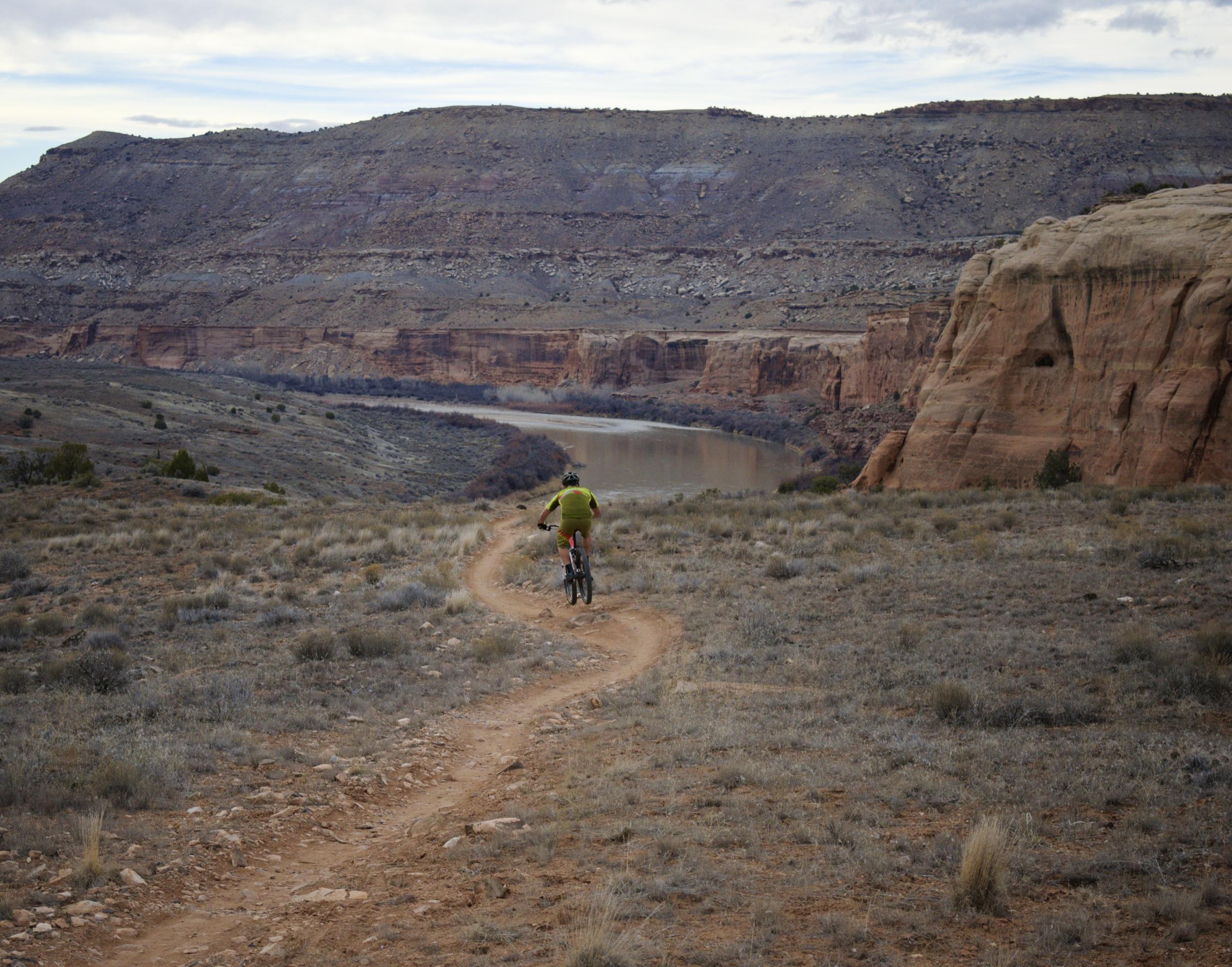 A mountain biker rides along a winding dirt trail overlooking a river, with rocky hills and a cloudy sky in the background. The landscape features sparse vegetation and rugged terrain, emphasizing the natural beauty of the outdoor environment. Mary's Loop / Horsethief Bench mountain bike trail.