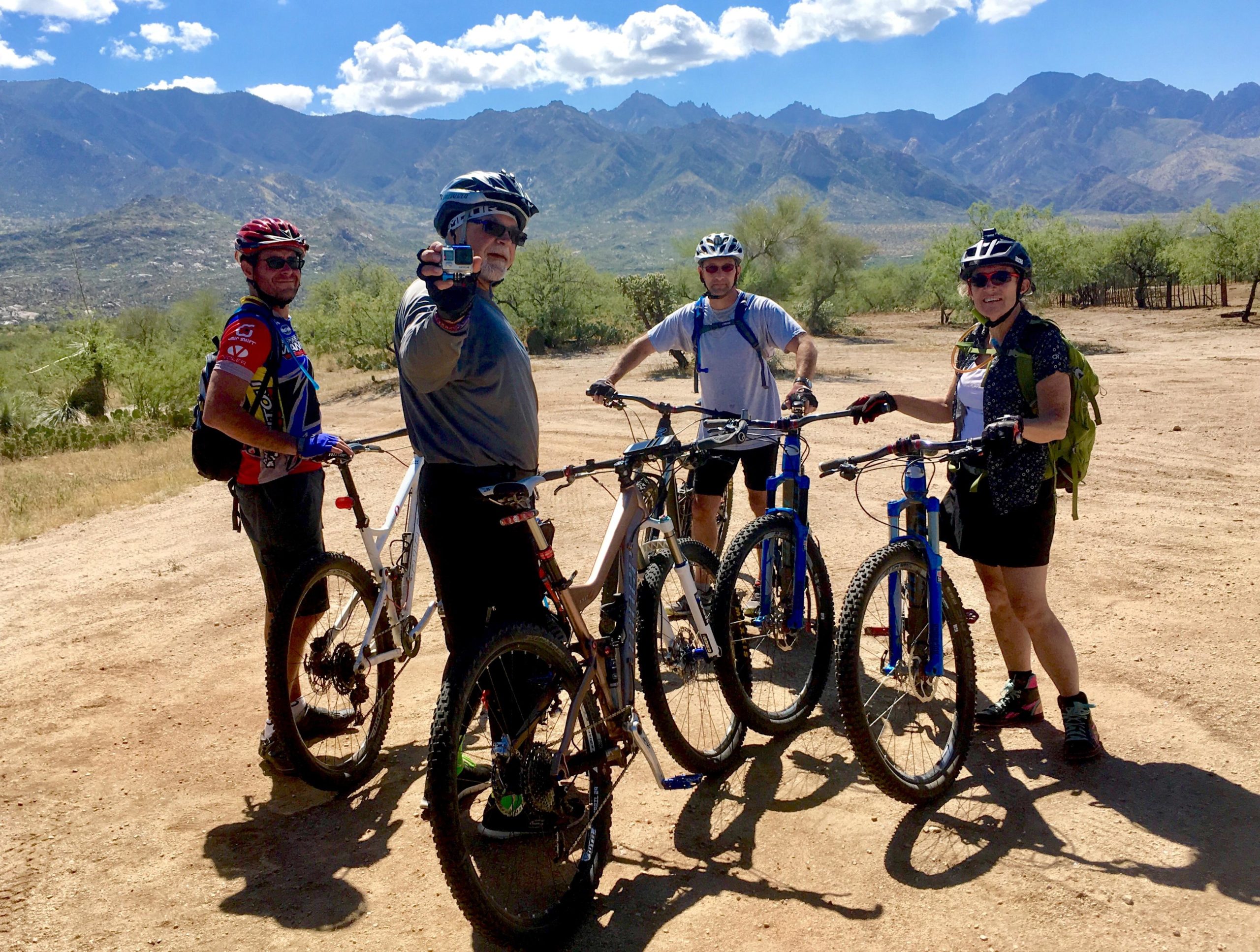 Four mountain bikers gather in a scenic outdoor setting, surrounded by mountains and blue skies. They are standing next to their bikes, with two men and two women, all wearing helmets and outdoor gear. One man is holding a camera, while the others are smiling and engaging in conversation. The terrain is dusty, indicating a recent ride in nature. 50-year Trail / Golder Ranch mountain bike trail.