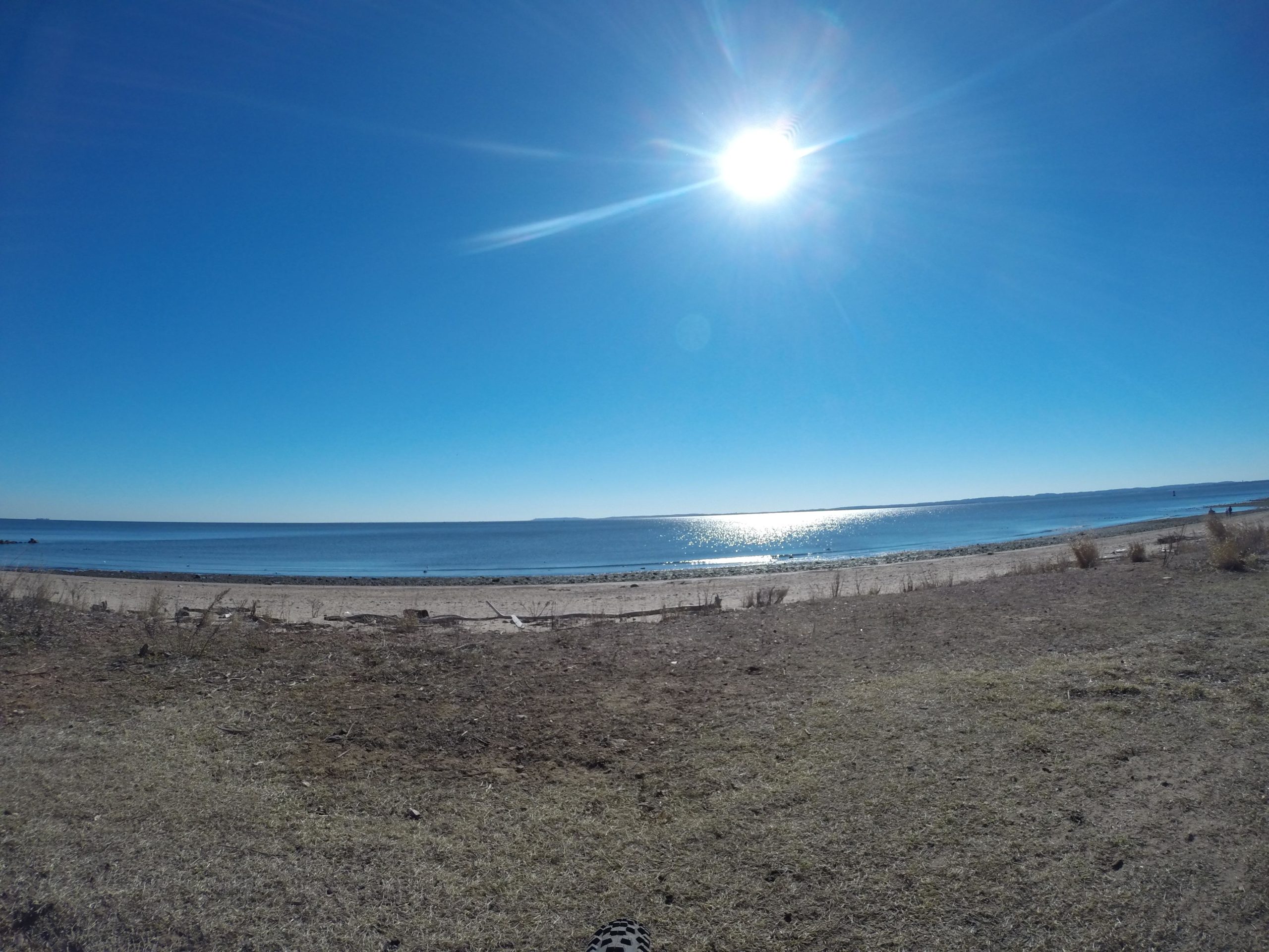 A serene beach scene under a clear blue sky. The sun shines brightly overhead, casting reflections on the calm water. The shoreline features sandy beach and patches of grass, with gentle waves lapping at the shore. Wolfes Pond park mountain bike trail.