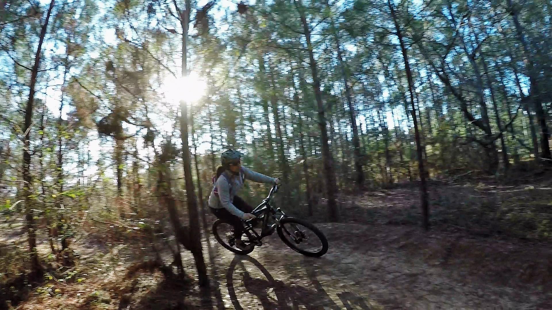 A person riding a mountain bike on a dirt trail through a forest, with the sun shining through the trees in the background. The rider is captured mid-motion, performing a stunt with the bike as they navigate the natural landscape. Beaver Creek / Forever Wild Trails mountain bike trail.