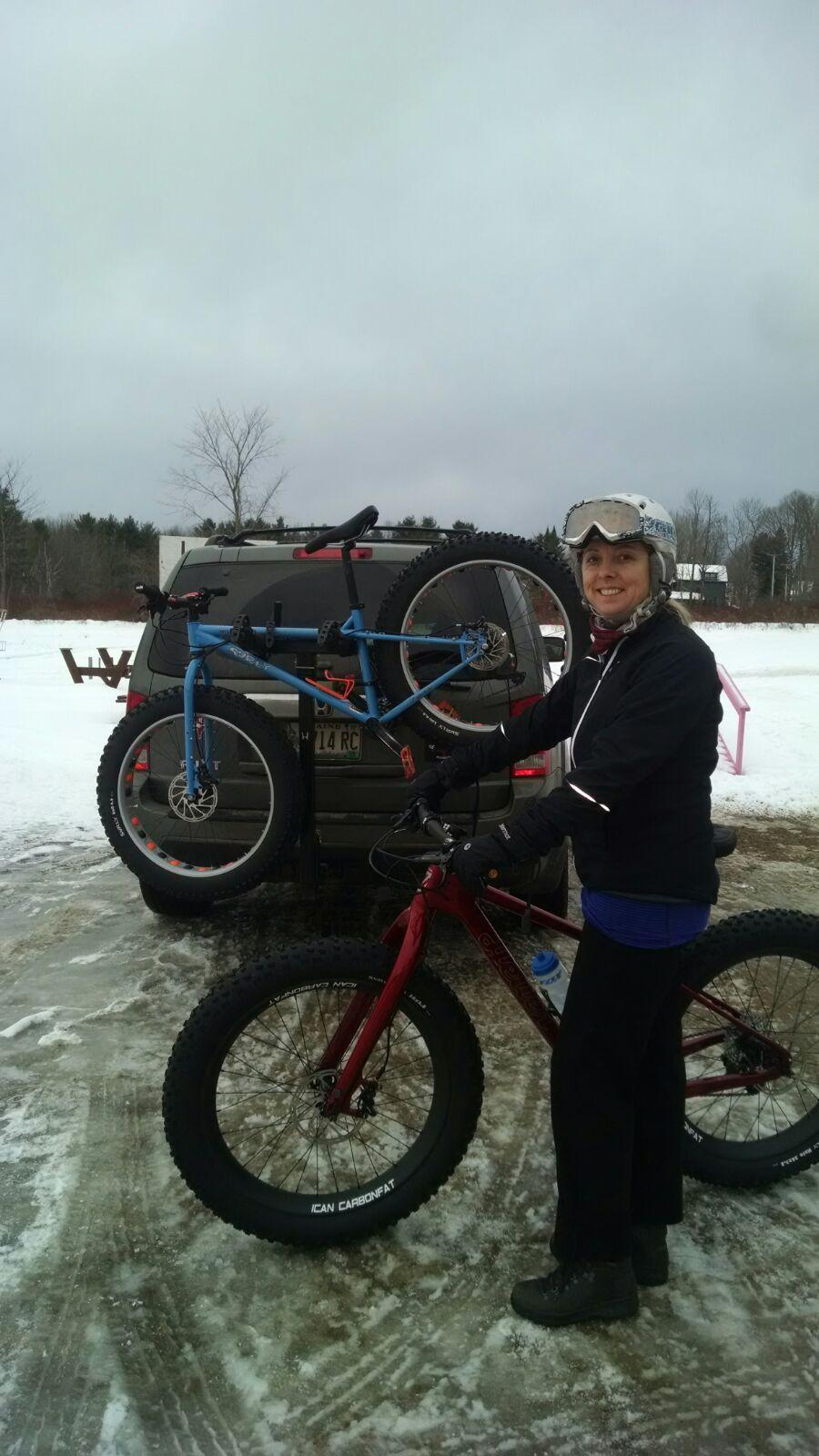 Heller Bloodhound: A person smiling beside a red fat bike parked on snow and ice, with a blue bike mounted on the back of an SUV in the background. The individual is wearing a helmet, gloves, and a black jacket, ready for biking in a winter setting. Overcast skies and trees are visible in the background.