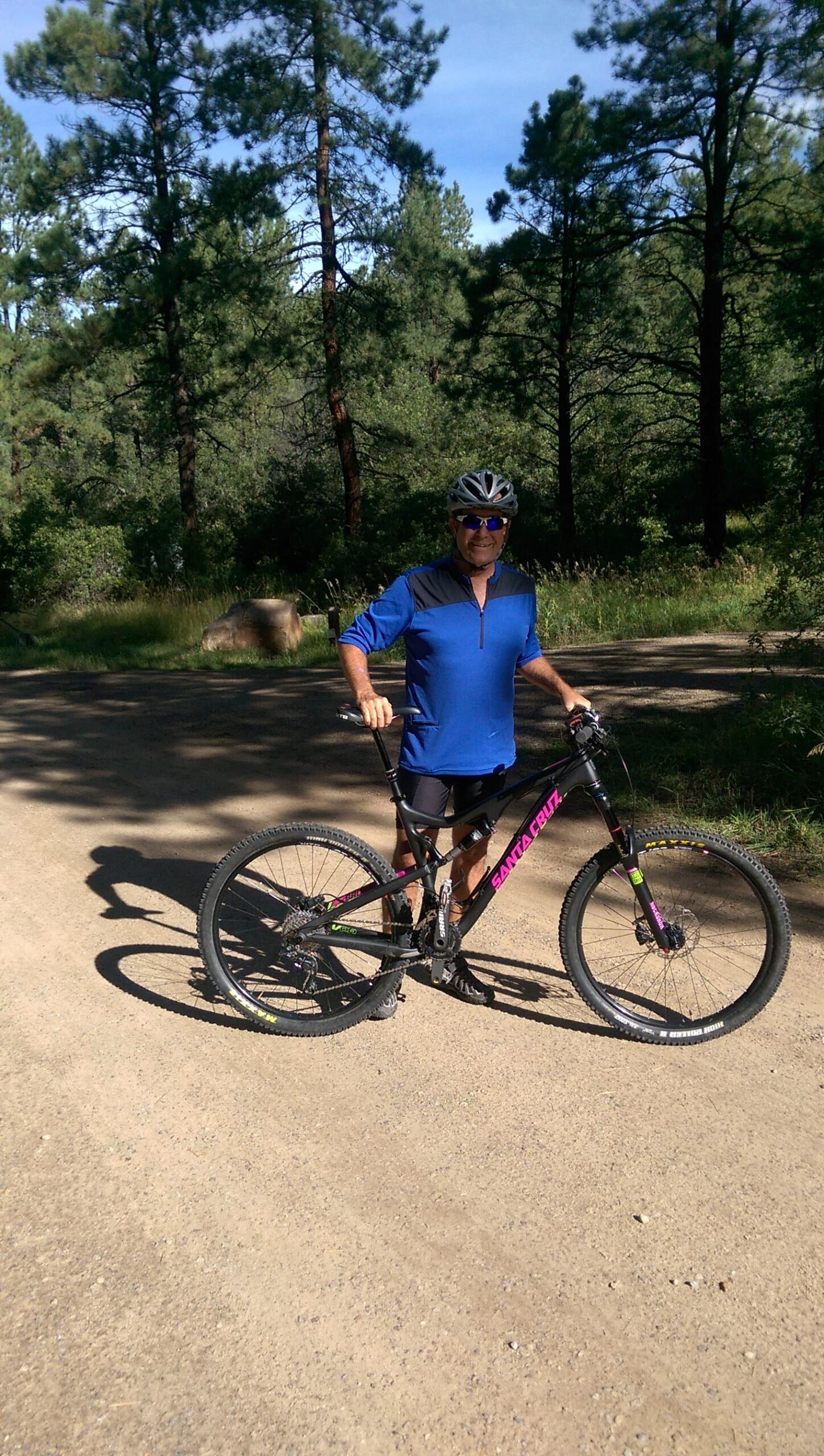 Santa Cruz Bronson: A cyclist in a blue long-sleeve shirt and black shorts stands next to a mountain bike in a natural setting. Tall pine trees and greenery surround a dirt path under a clear blue sky. The cyclist is wearing a helmet and sunglasses, appearing ready for a ride in the outdoors.