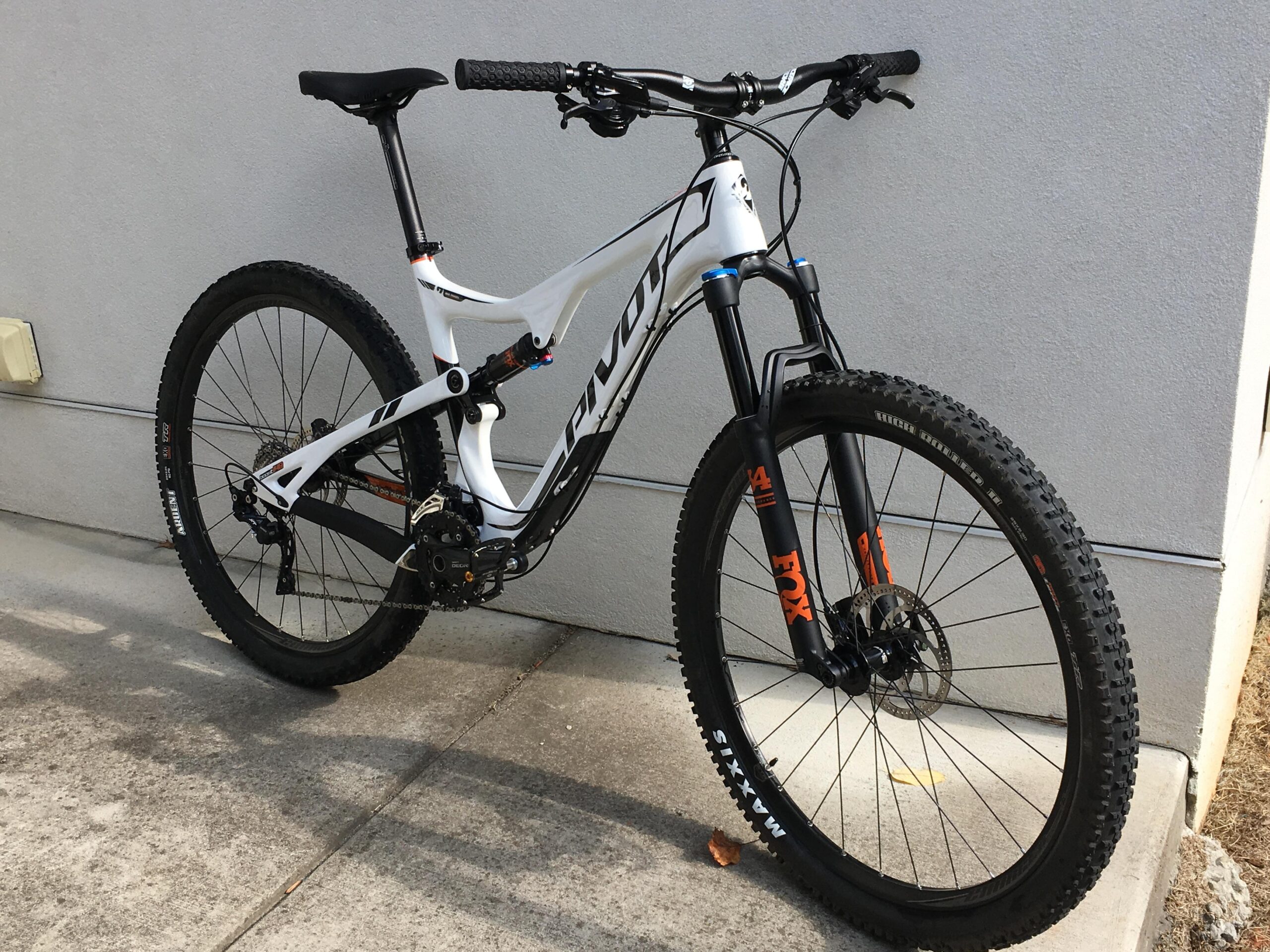 Pivot Trail 429: A white and black mountain bike with a sturdy frame, equipped with knobby tires and a front suspension fork. The bike is positioned on a concrete surface next to a textured gray wall, showcasing its modern design and components.