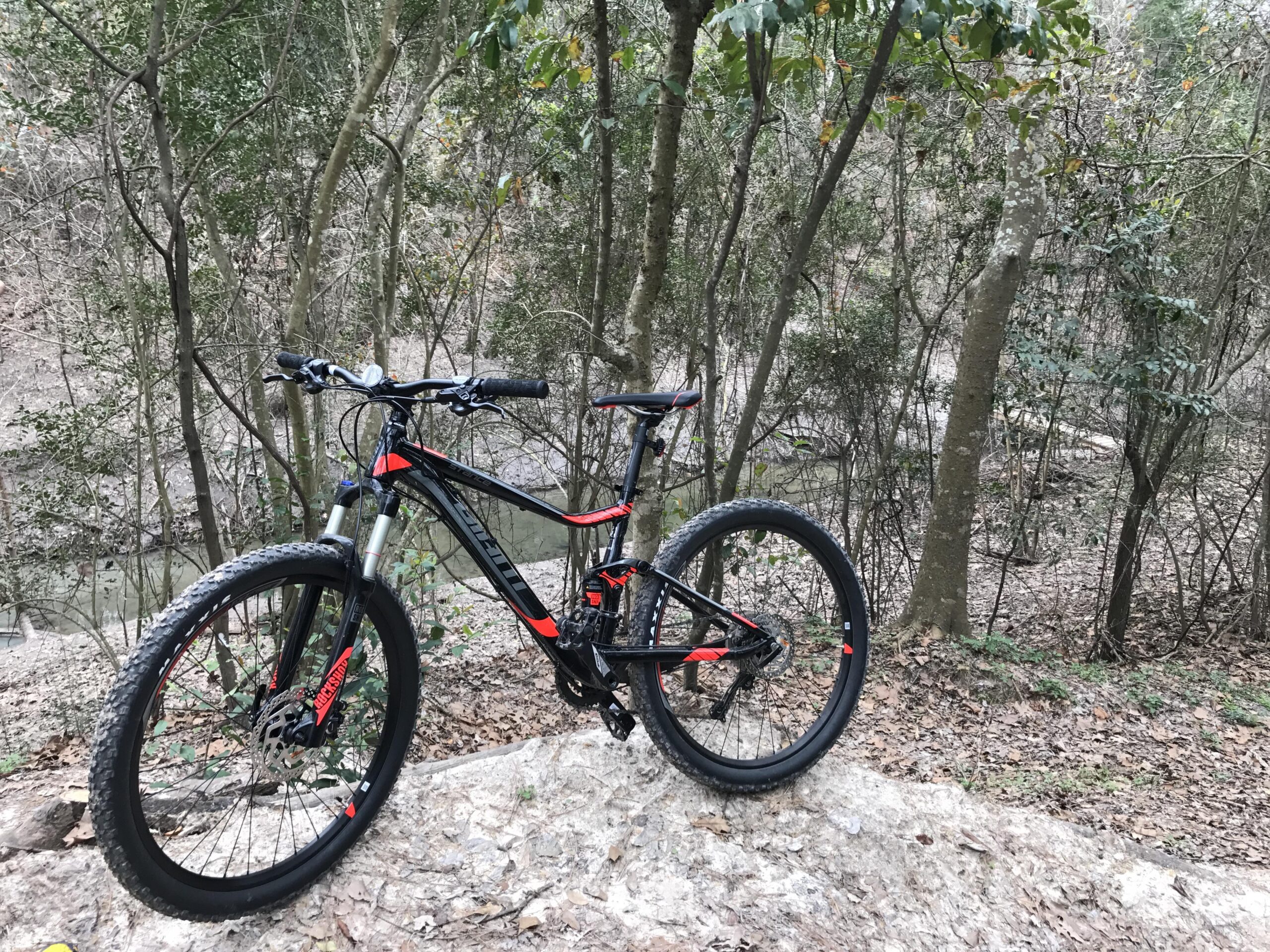 Giant Stance 27.5 2: A mountain bike resting on a rocky outcrop in a wooded area, surrounded by trees and foliage, with a clear view of a stream in the background.