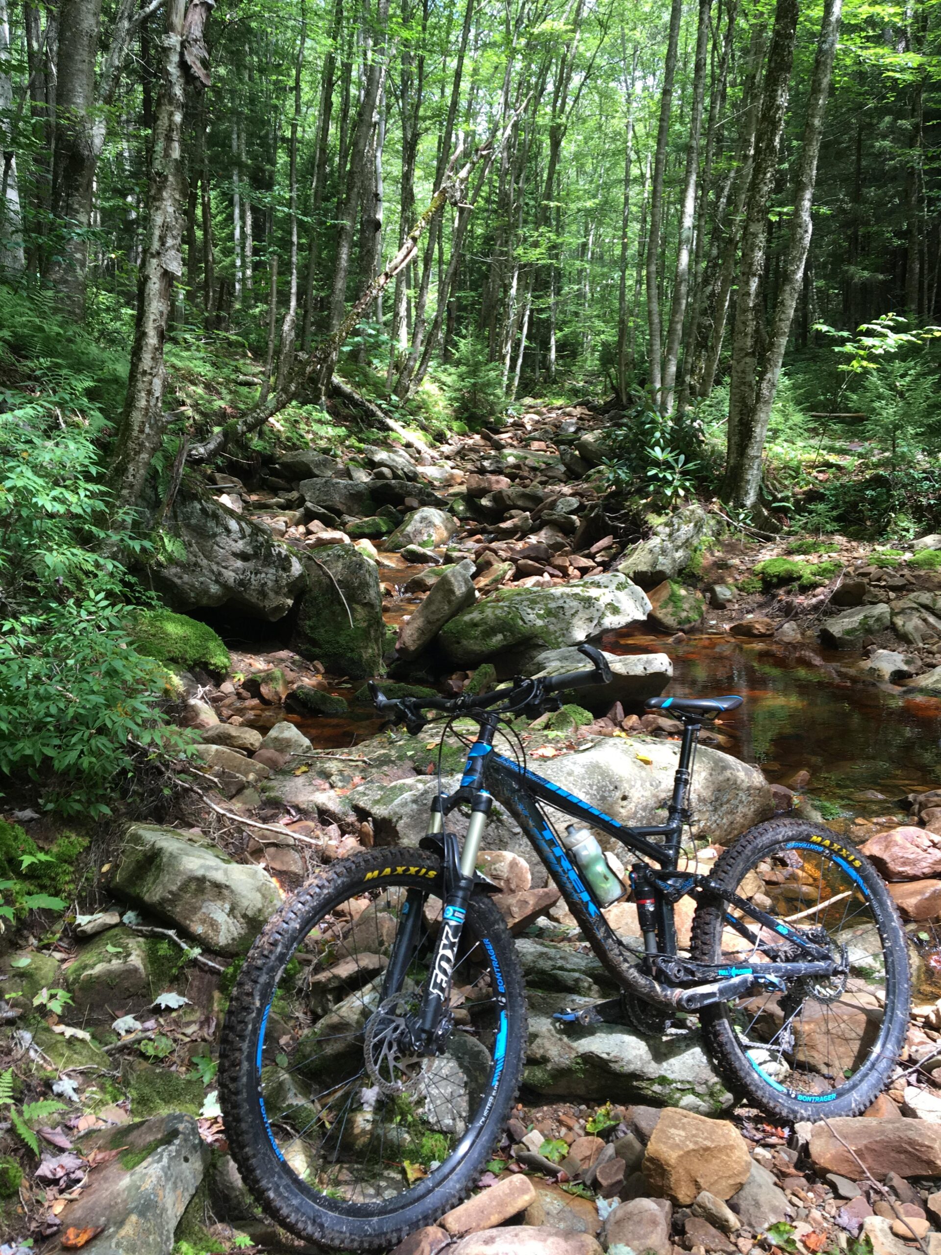Trek 2014 trek fuel ex 8 27.5: A mountain bike leaning against a large rock by a rocky stream in a lush green forest. Sunlight filters through the trees, creating a serene outdoor scene.