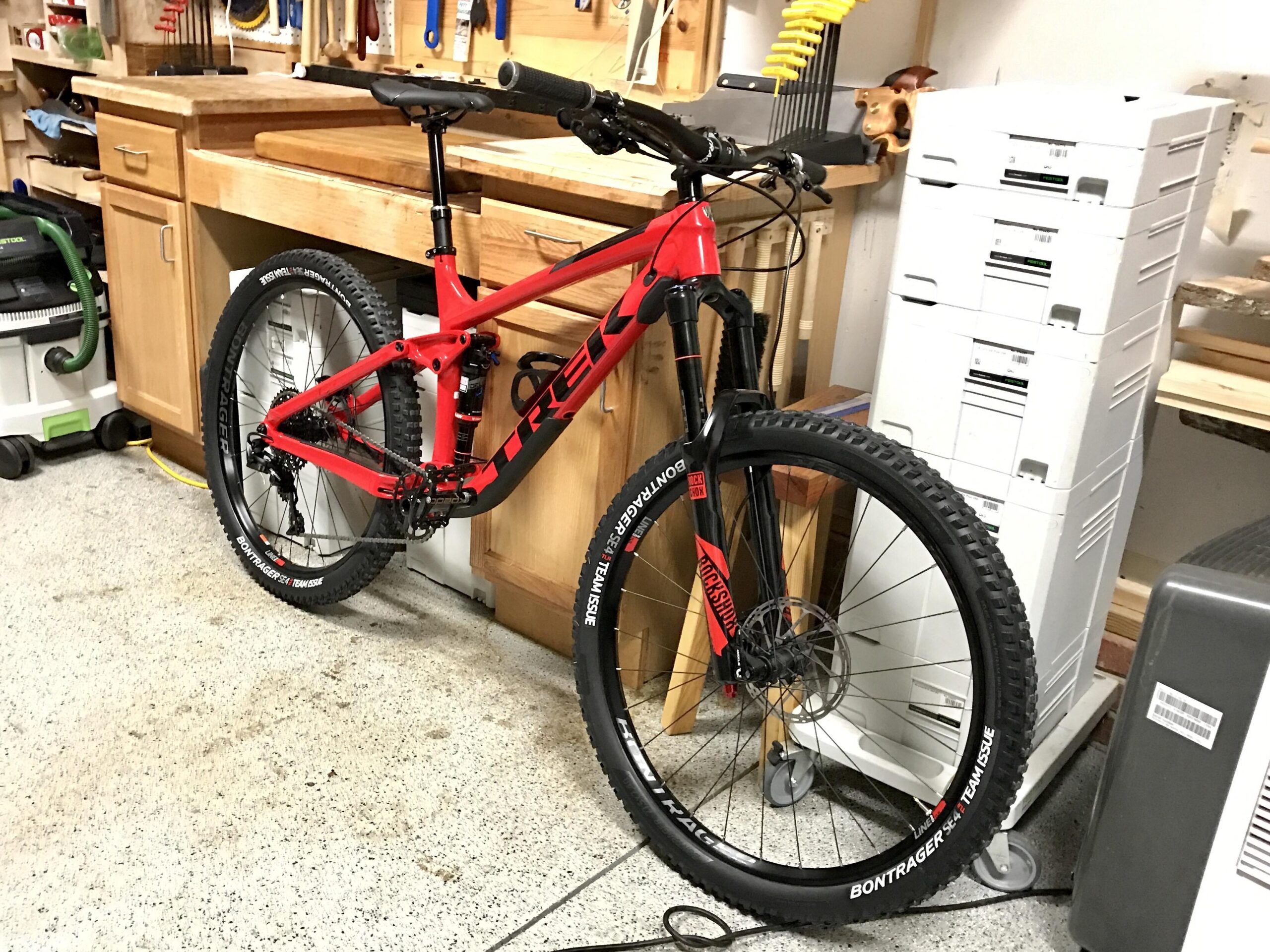Trek Remedy 9: A red Trek mountain bike is parked beside a wooden workbench in a garage setting. The bike has thick, knobby tires and features components typical of a high-performance mountain bike. In the background, various tools and equipment are visible on shelves, along with storage units and a vacuum cleaner. The garage floor shows a bit of wear, indicating practical use.