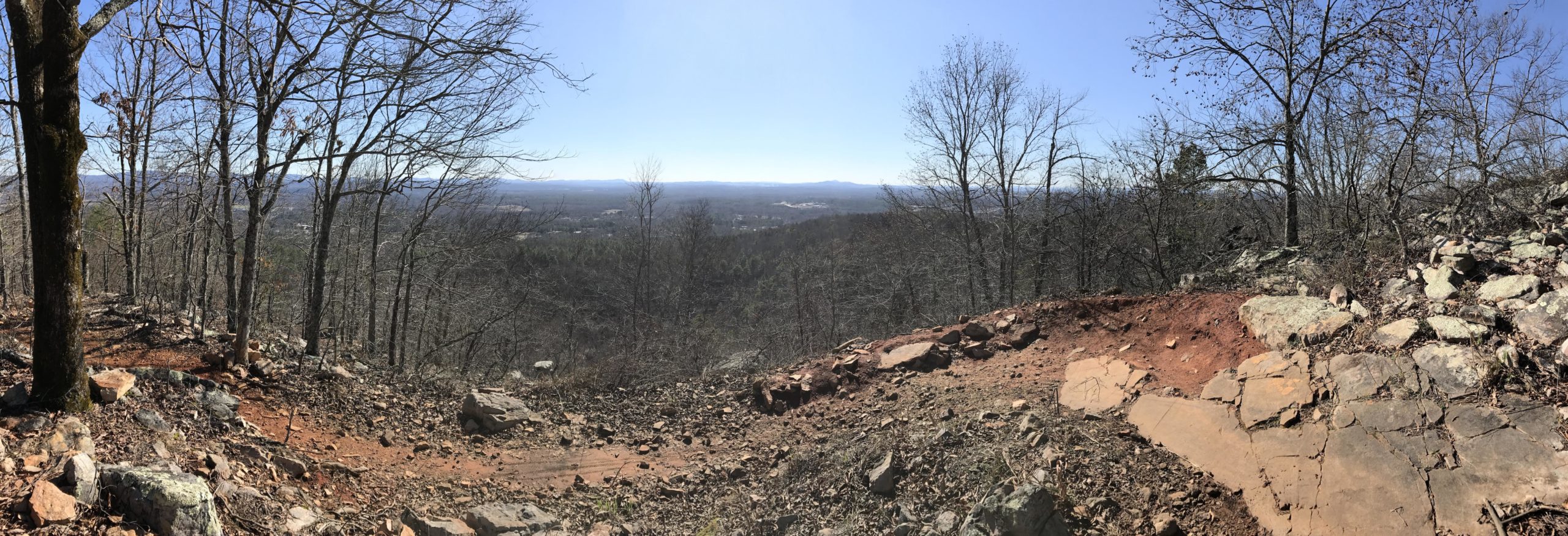 A panoramic view of a hilly landscape during a clear day, featuring sparse trees with no leaves, rocky terrain, and an expansive sky. The foreground includes scattered rocks and a dirt path leading to the horizon, while the background offers a distant view of rolling hills and valleys. Coldwater Mountain mountain bike trail.