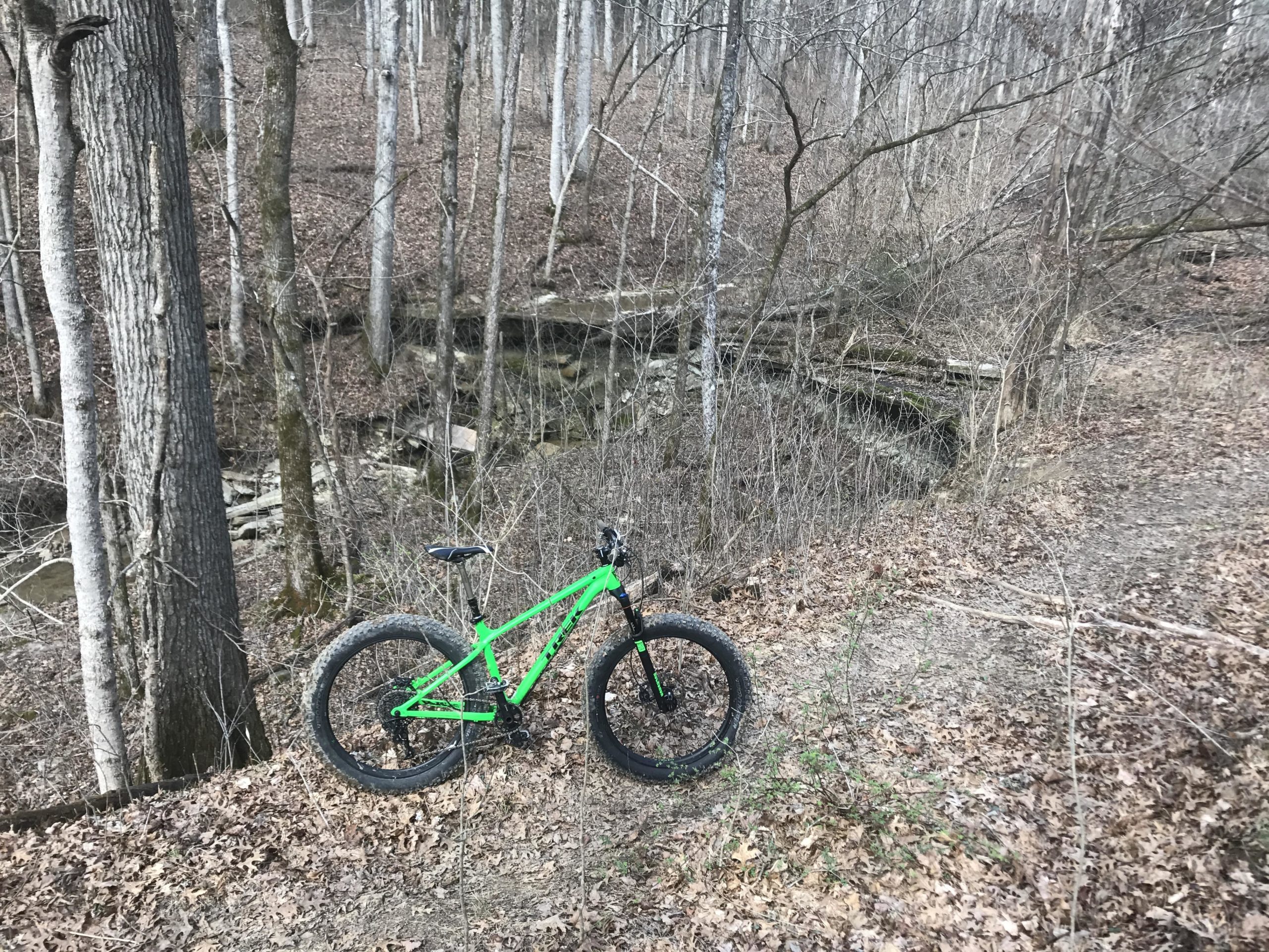 A bright green mountain bike parked on a dirt trail surrounded by bare trees and underbrush in a forested area. In the background, a shallow creek is visible, along with rocky terrain and fallen branches. The setting appears calm and natural, suggesting a remote outdoor location suitable for biking and exploring. Karst climb mountain bike trail.
