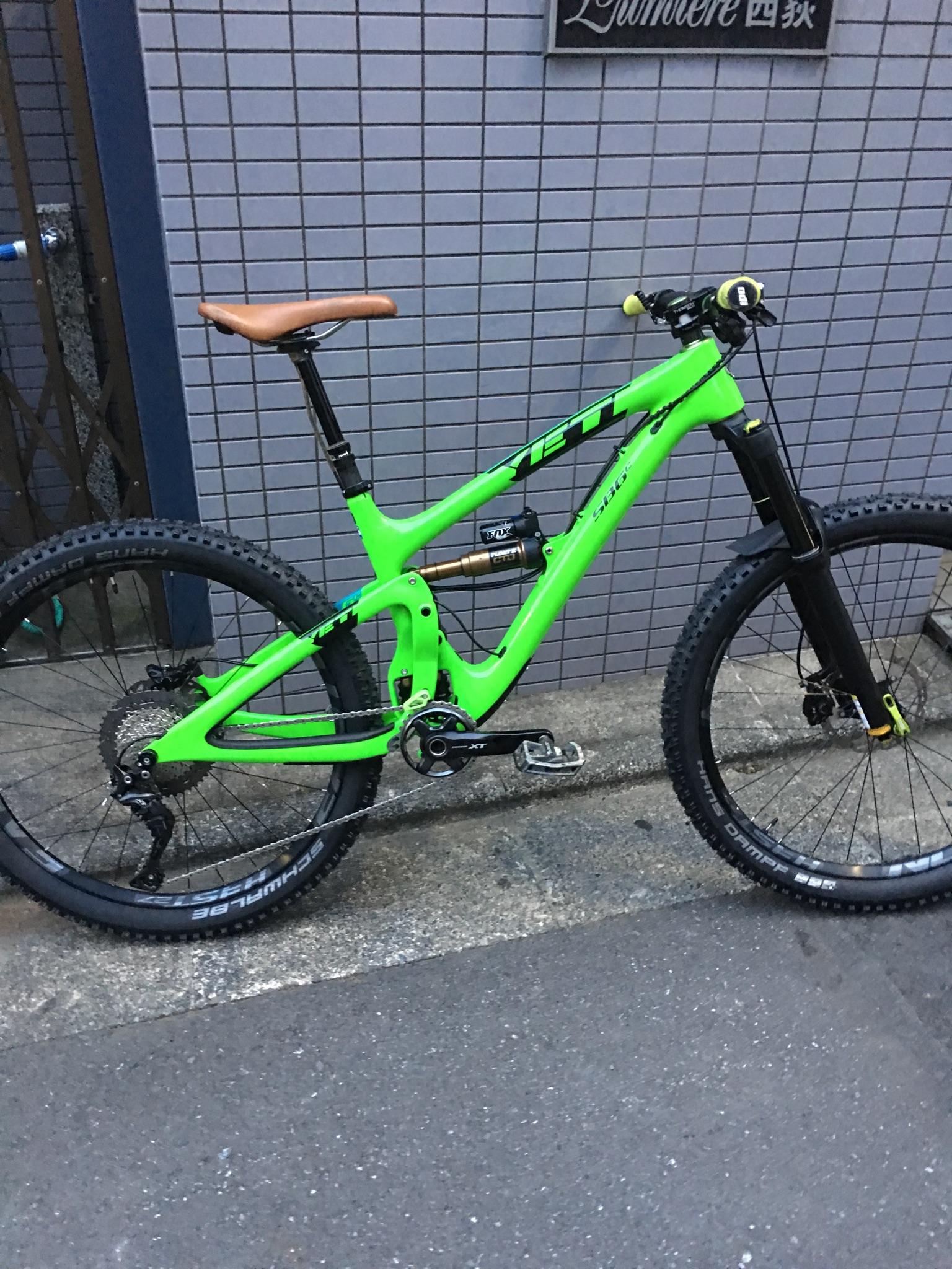 Yeti SB6: A bright green mountain bike with a brown leather seat, parked on a concrete surface next to a textured gray wall. The bike features rugged tires and a front suspension fork, emphasizing its suitability for off-road terrain.