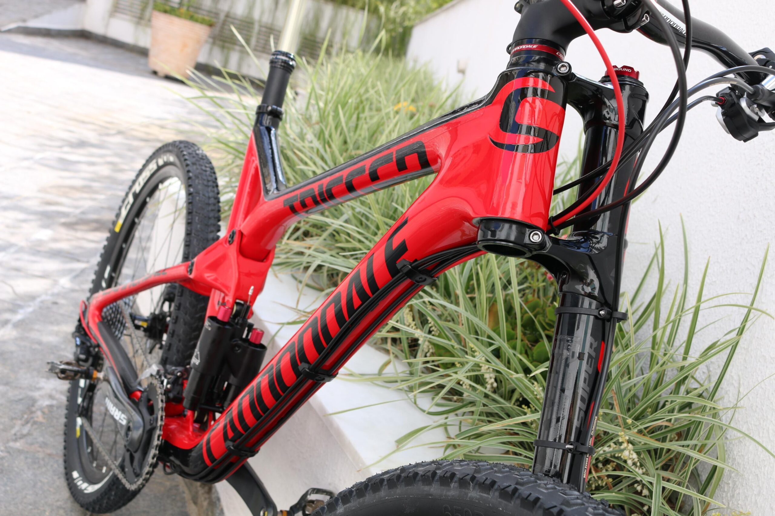 Cannondale Trigger Carbon 2 27.5: Alt text: A close-up view of a red mountain bike frame with black accents, featuring a full suspension design and a prominent logo. The bike is positioned on a paved surface with green landscaping in the background.