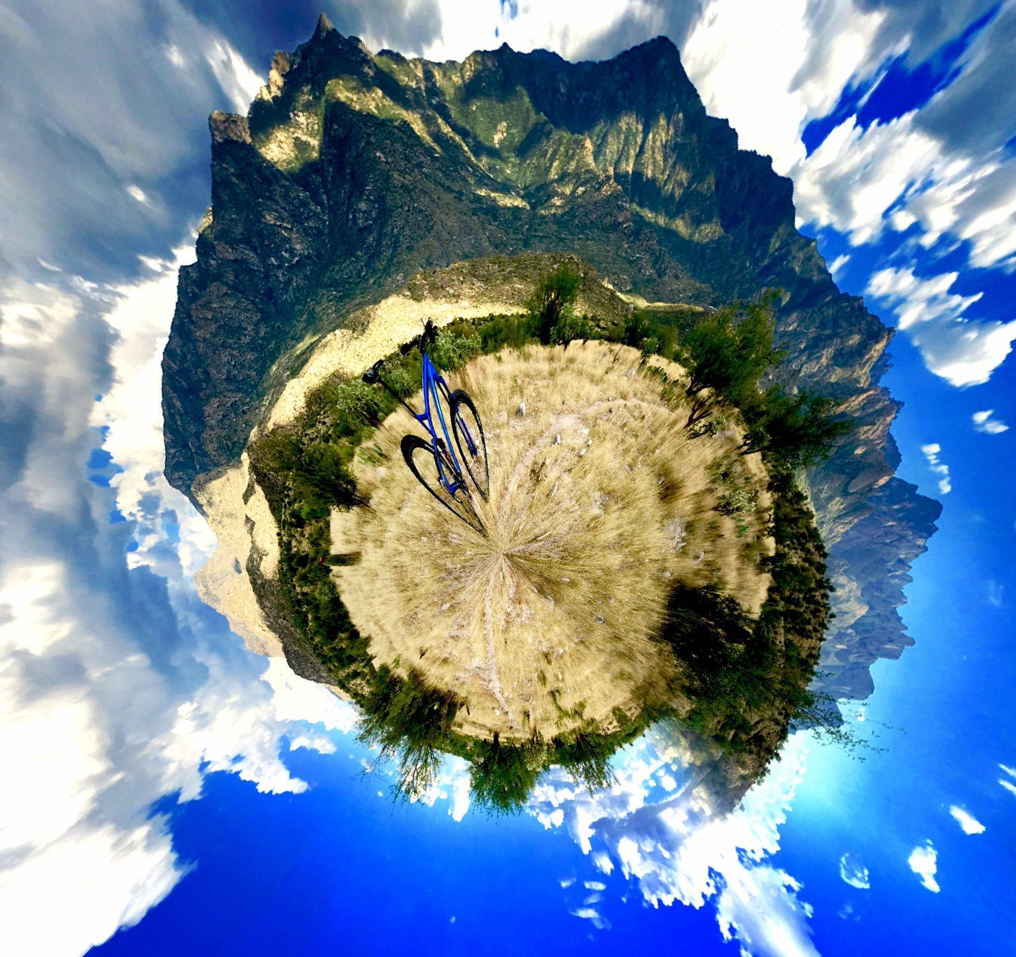 A vibrant panoramic view of a mountainous landscape, featuring a blue mountain bike in the foreground. The scene is characterized by a surreal, spherical perspective, with dramatic clouds and bright blue sky enhancing the natural beauty. Lush green vegetation and dry terrain are visible, creating a striking contrast with the majestic mountains in the background. 50-year Trail / Golder Ranch mountain bike trail.