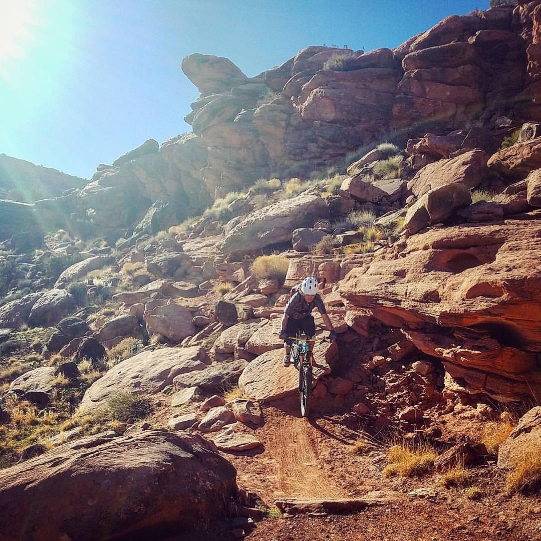 A mountain biker navigating a rocky trail in a desert landscape, surrounded by large boulders and sparse vegetation under a clear blue sky. The sun is shining brightly, casting warm light over the scene. Captain Ahab mountain bike trail.