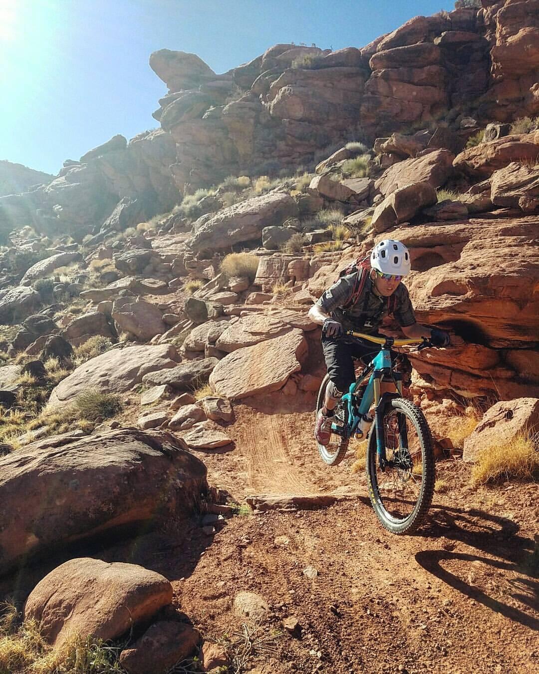 A cyclist wearing a helmet and protective gear rides a mountain bike along a rocky trail in a desert landscape. Sunlight illuminates the scene, highlighting the rugged terrain and scattered vegetation. Captain Ahab mountain bike trail.