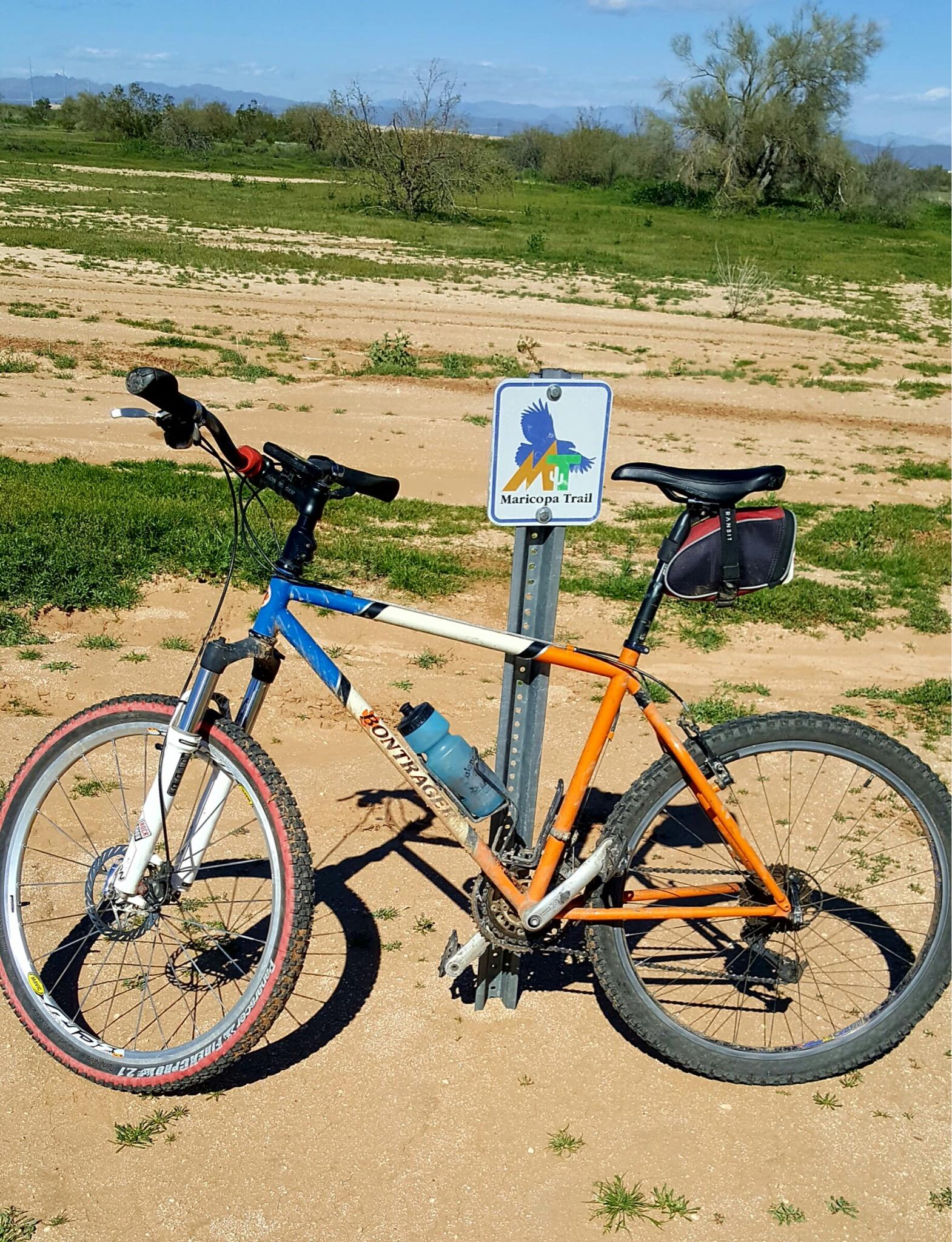 A mountain bike with blue and orange frame colors parked next to a sign for the Maricopa Trail, set in a sandy area with sparse vegetation and distant hills in the background. White Tanks Regional Park Trails mountain bike trail.