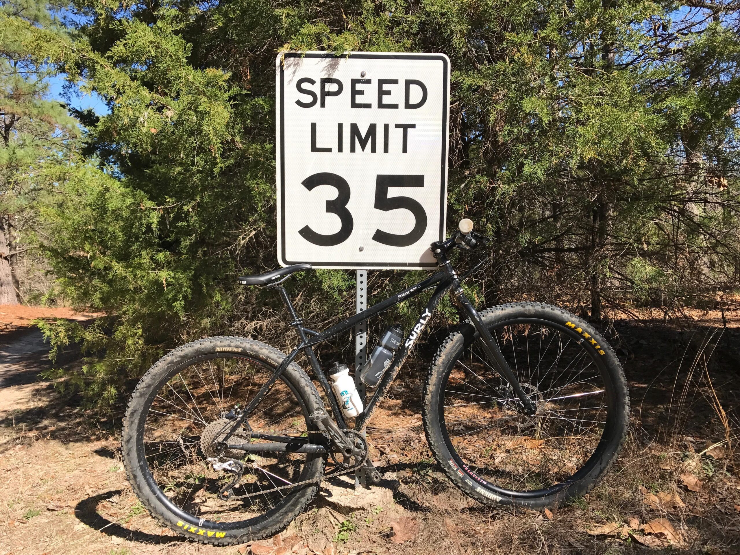 Surly Karate Monkey: A black bicycle leaning against a speed limit sign that reads "35" in a wooded area. The background features green trees and a dirt path, suggesting a rural or natural setting.